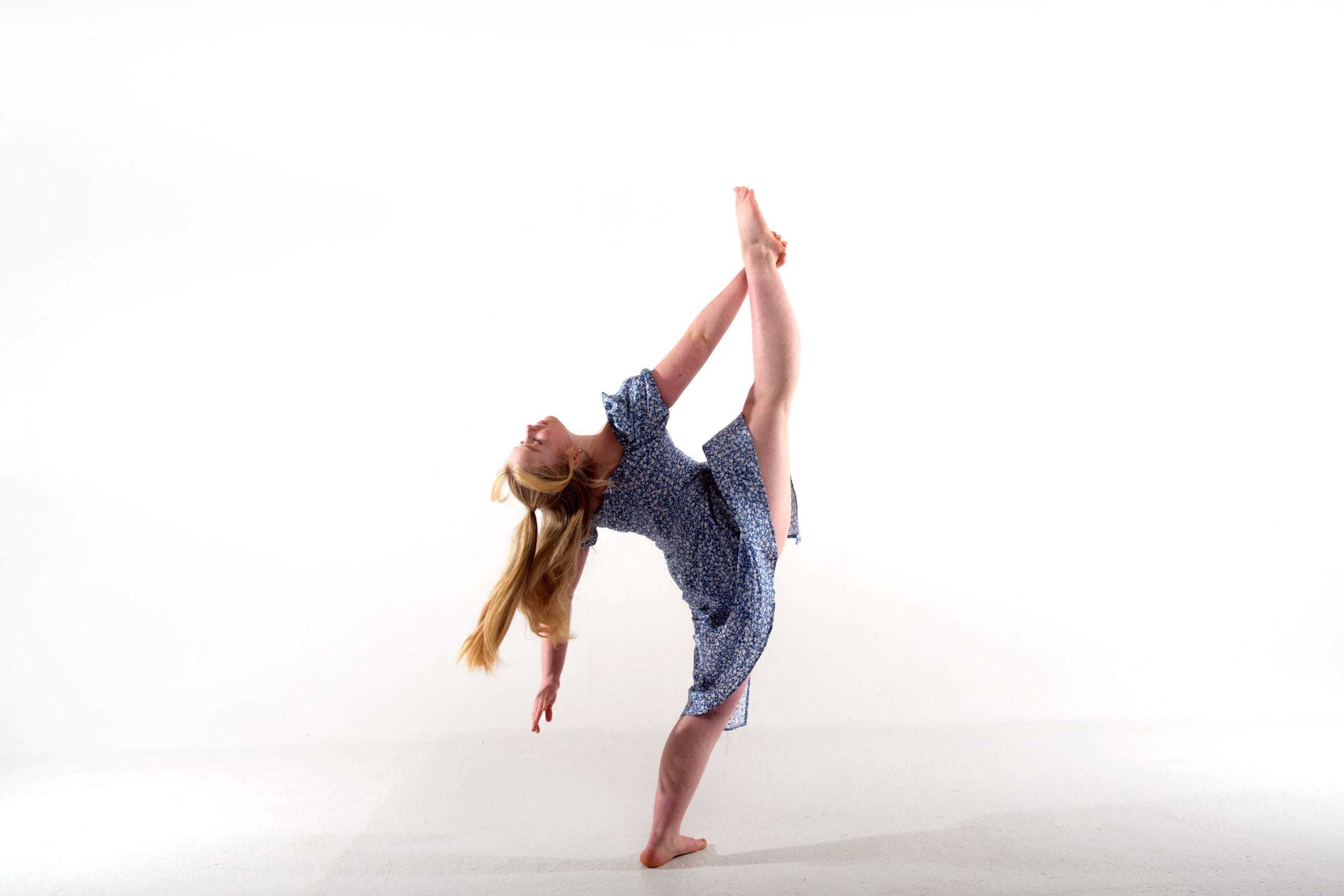 Against a white background, a female dancer is photographed in a standing straddle pose with pointed feet. She has long blonde hair and is wearing a blue dress.