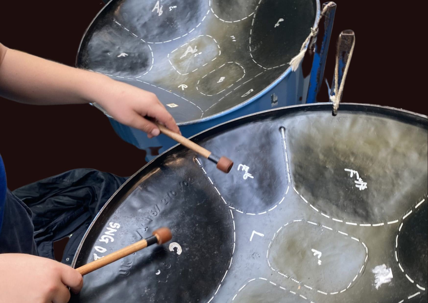 A close up of two steel pans, they are being played by a person whose hands are visible holding wooden sticks.