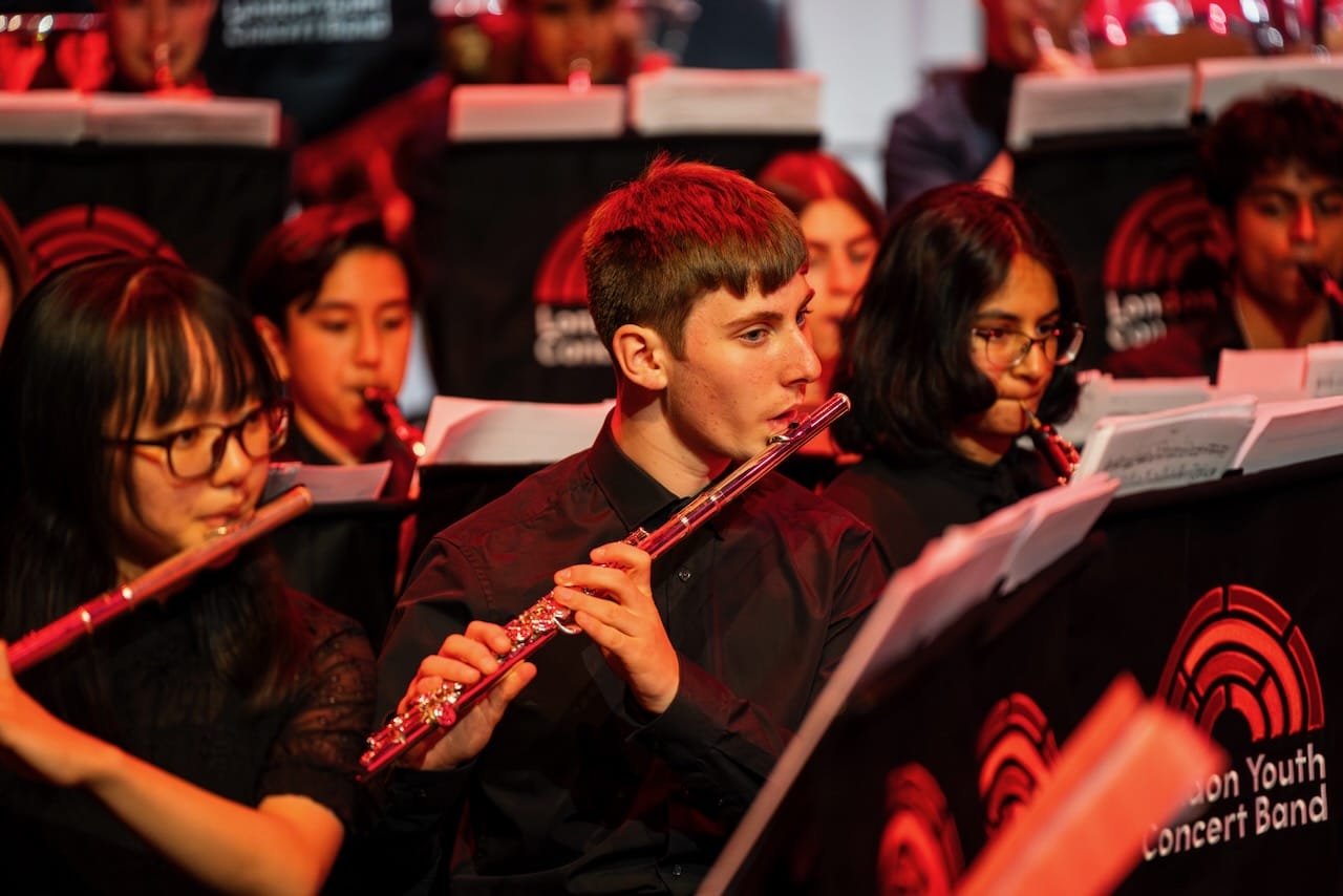 A professional photograph of London Youth Concert Band in performance. We see three featured musicians playing flute and other wind instruments. The image is illuminated by red stage lighting.