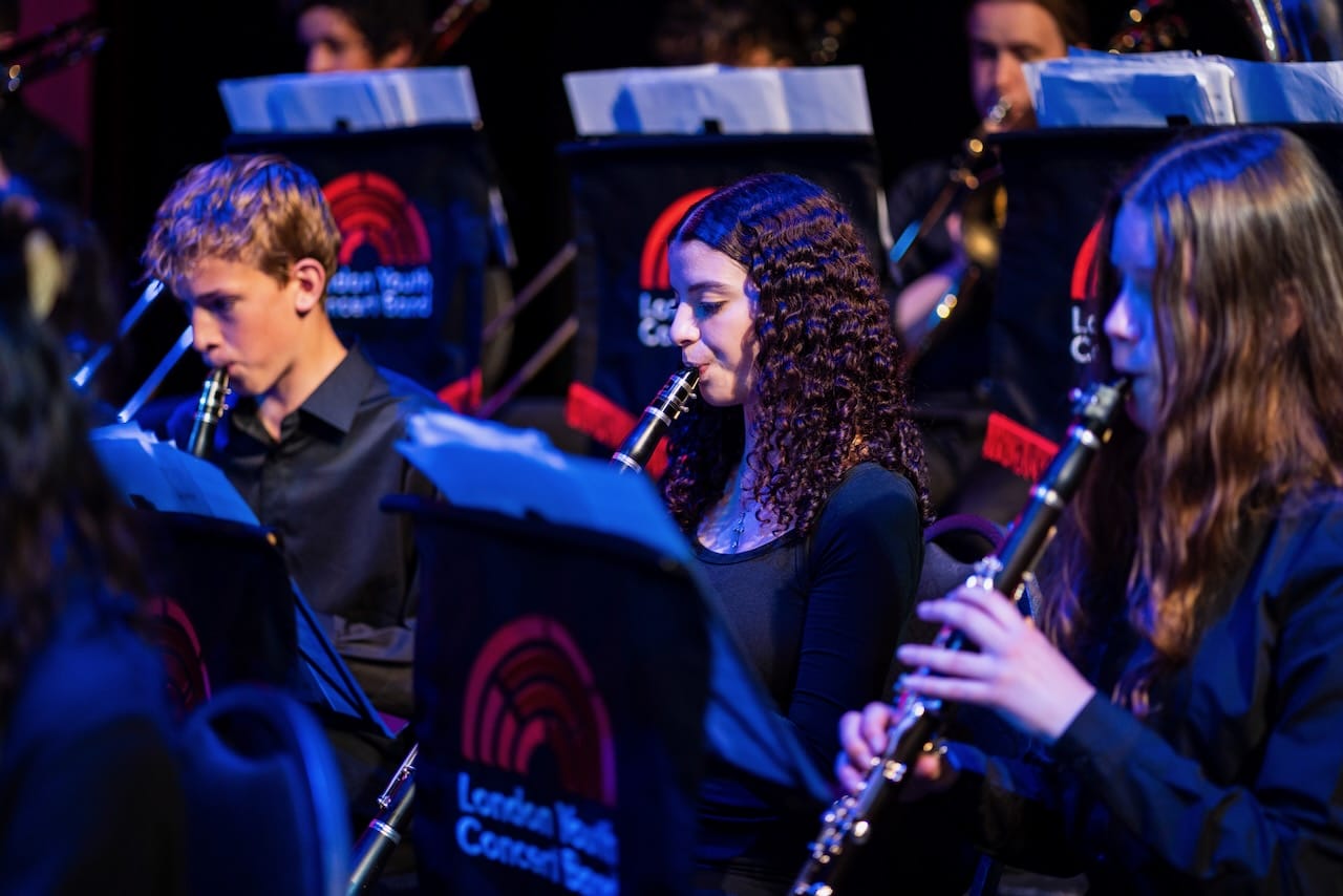 Close up photograph of London Youth Concert band members playing clarinet and other wind instruments. The photo has blue stage lighting illuminating the performers.