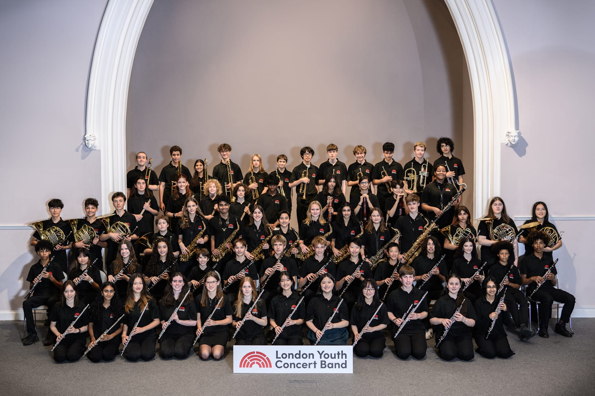 A full company photograph of the London Youth Concert band. Each member holds their instrument angled to the right. They wear uniform black shirts, and are posed underneath a large white arch.