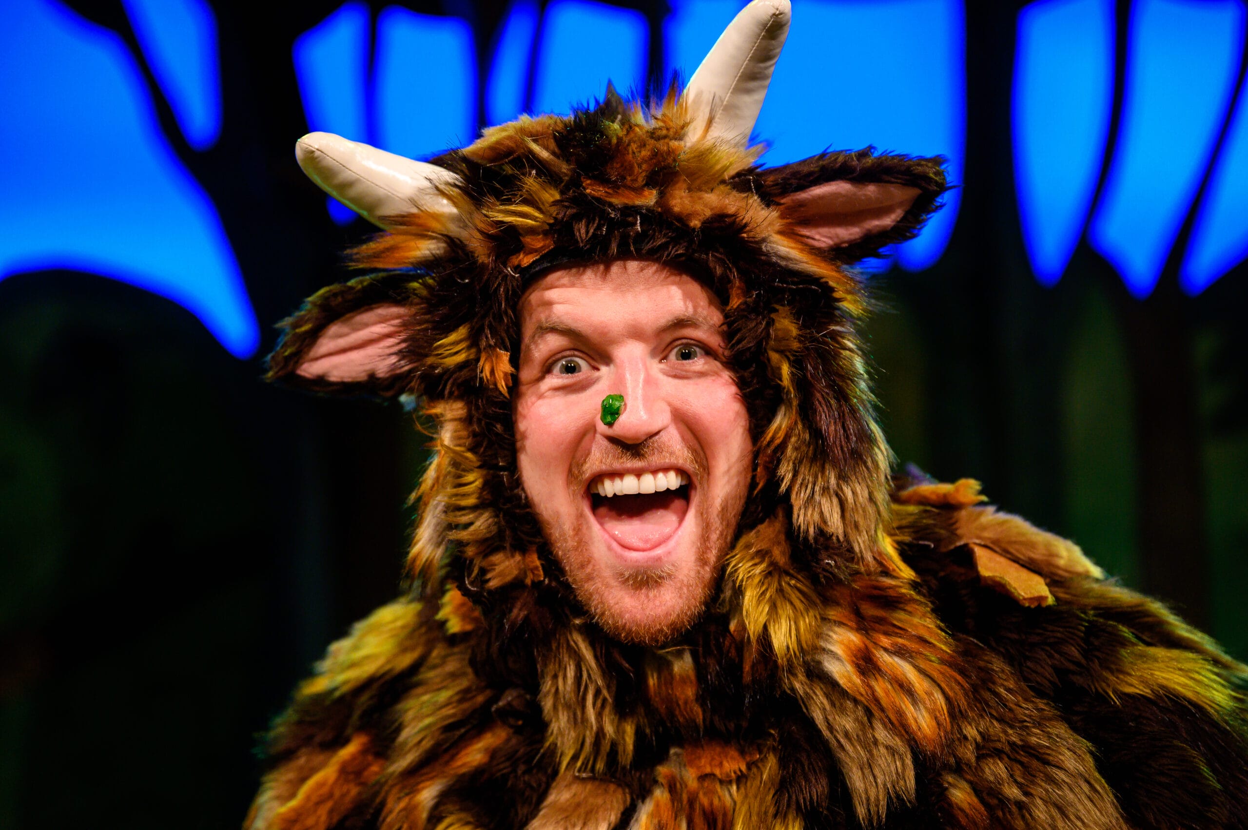 The Gruffalo actor wears a large fluffy costume with textured brown and amber fur, pink ears and large horns. He has an exaggerated green wart on his nose and is looking straight at the camera with a wide mouthed, excited expression.