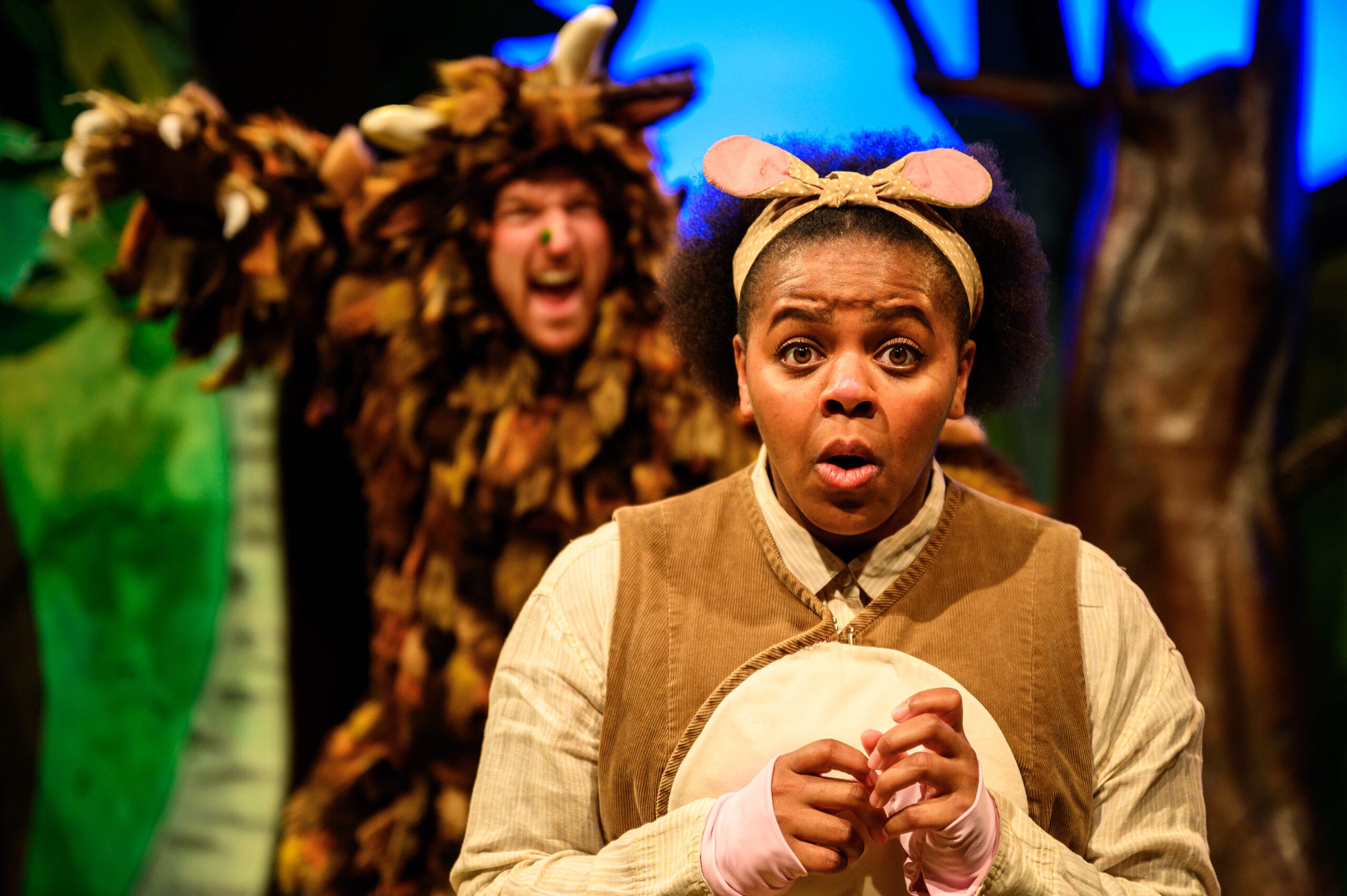 Mouse performer is centre stage. She wears a fawn coloured headband tied in a bow to look like mouse ears, accompanied by a brown and beige costume. She has an anxious expression and hands clasped together, whilst the Gruffalo creeps up behind her in the background of the image.