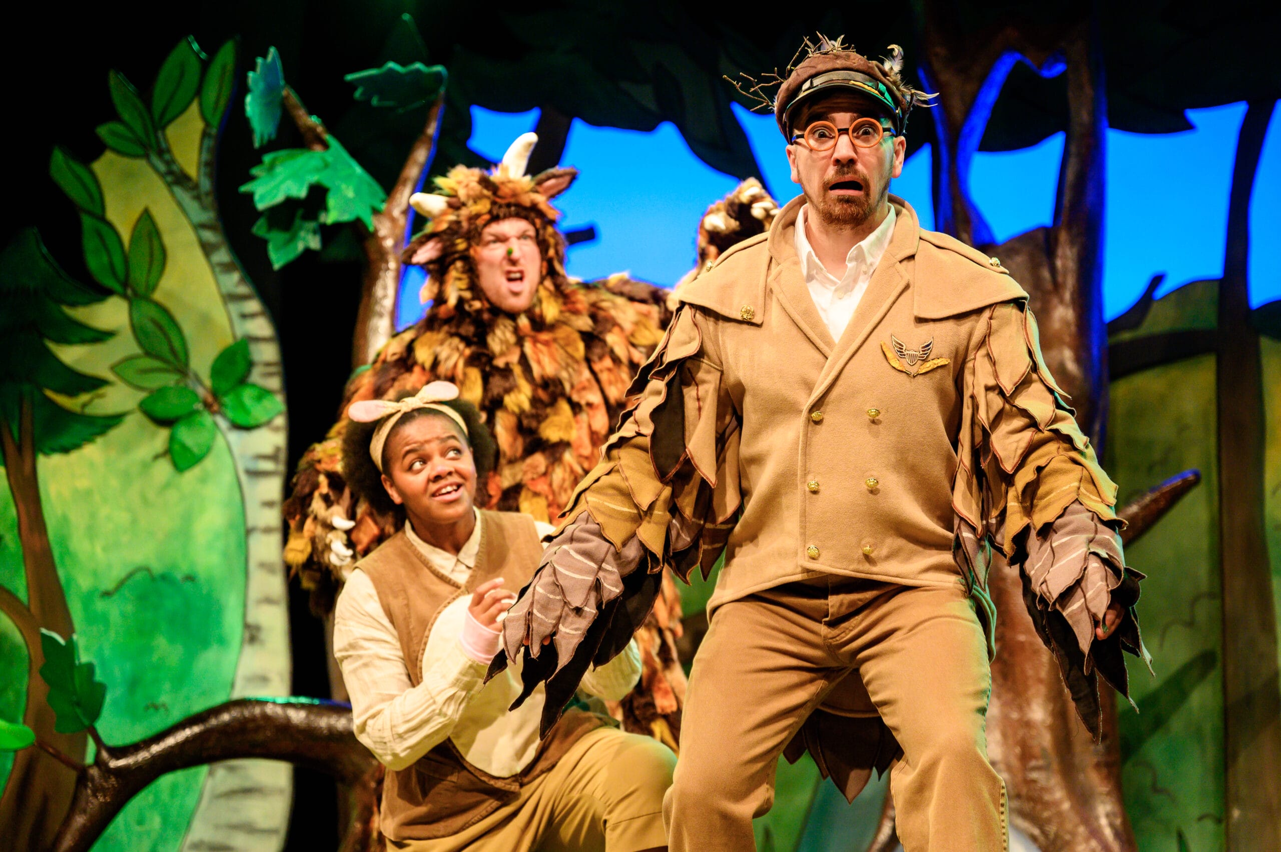The Gruffalo and Mouse are on the left side of the stage, looking up at Owl! Owl wears a beige suit with large wings on both arms and circle spectacles. Behind the actors are leafy green trees.