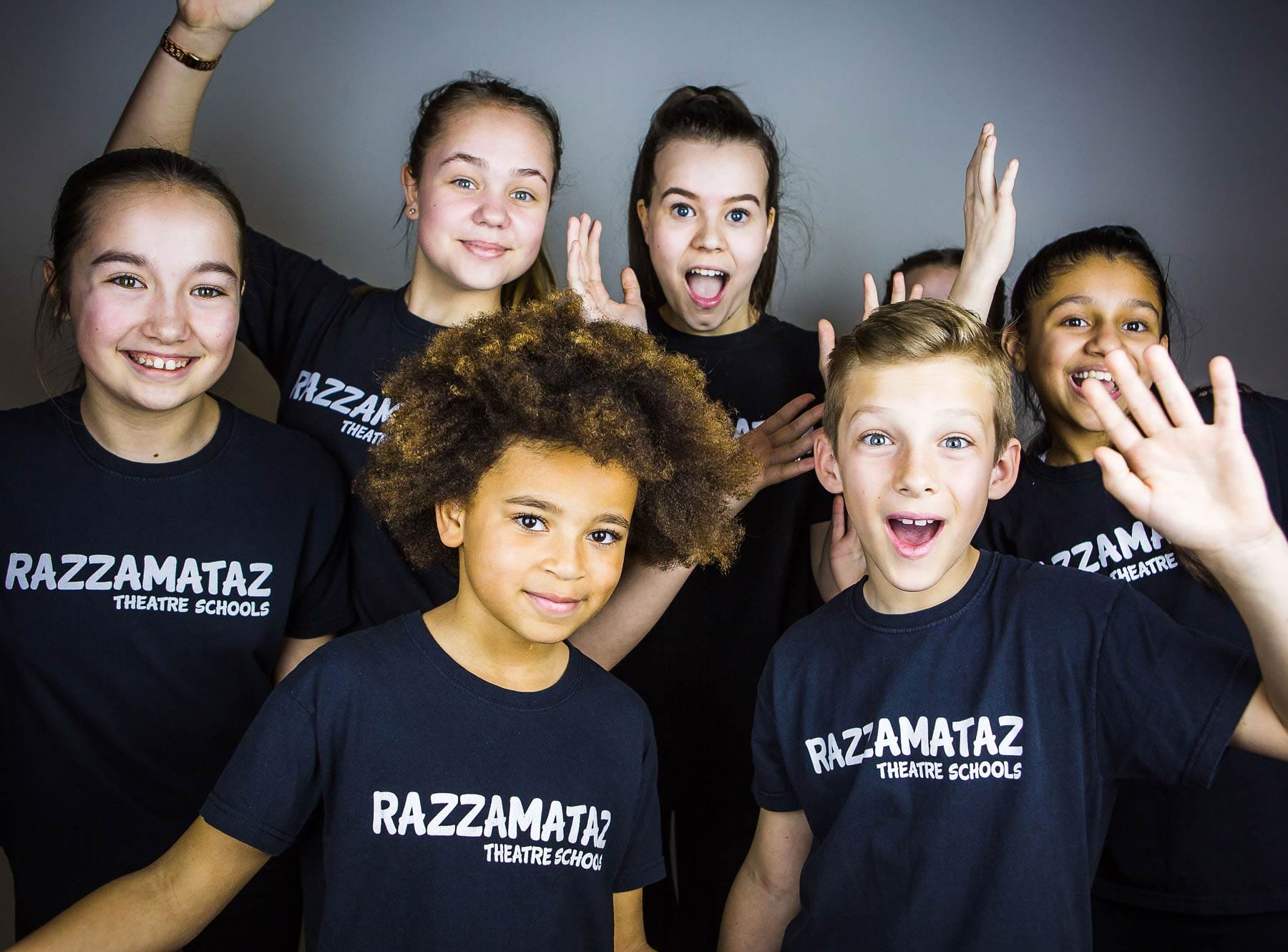 A group of young people striking dynamic poses, all wearing black t-shirts with white text reading RAZZAMATAZ Theatre Schools