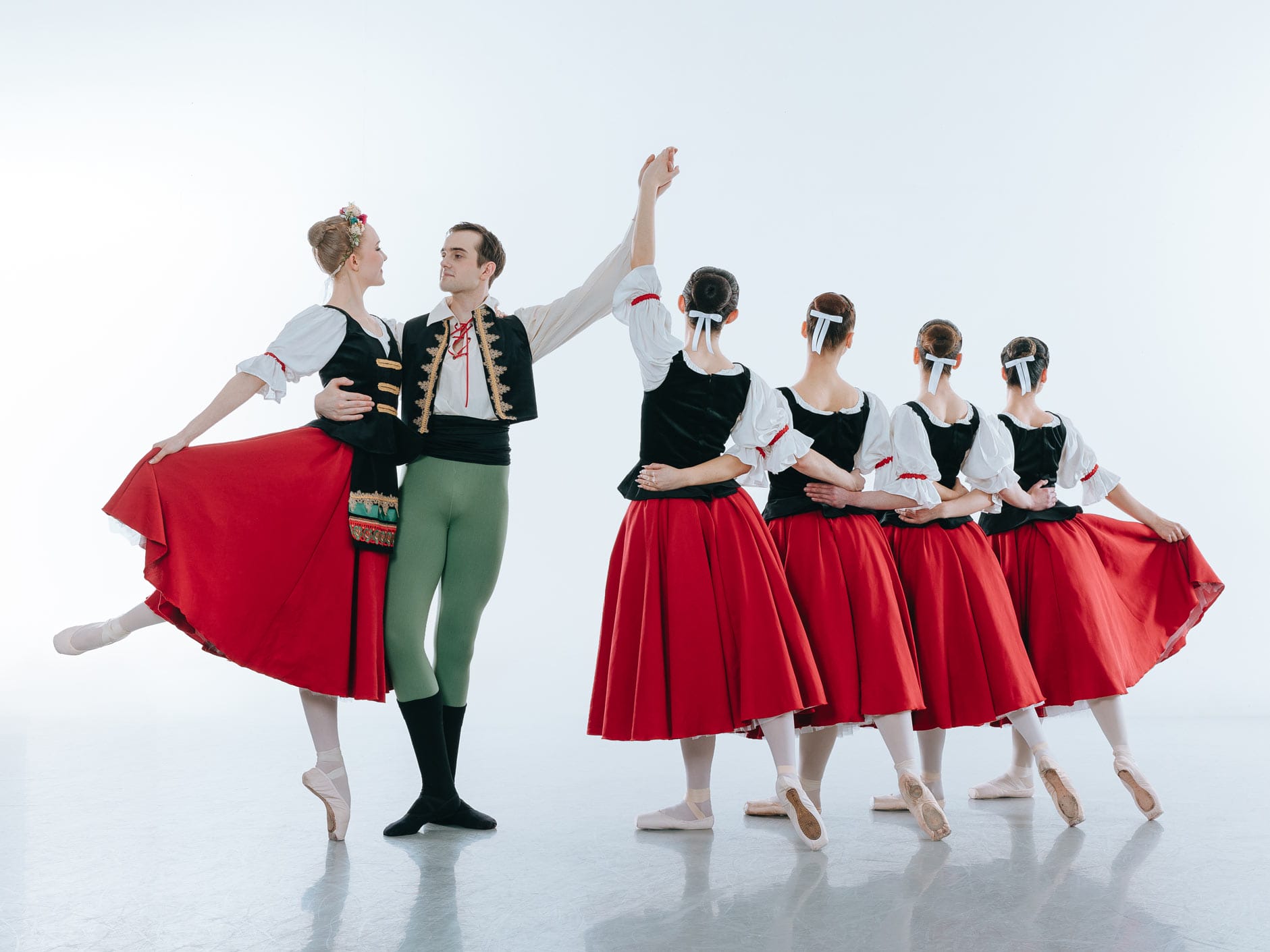 A group of 6 dancers wearing traditional ballet outfits. four female dancers stand with their back to the camera and their arms around each others waists. A female dancer balances en pointe supported by a male dancer.