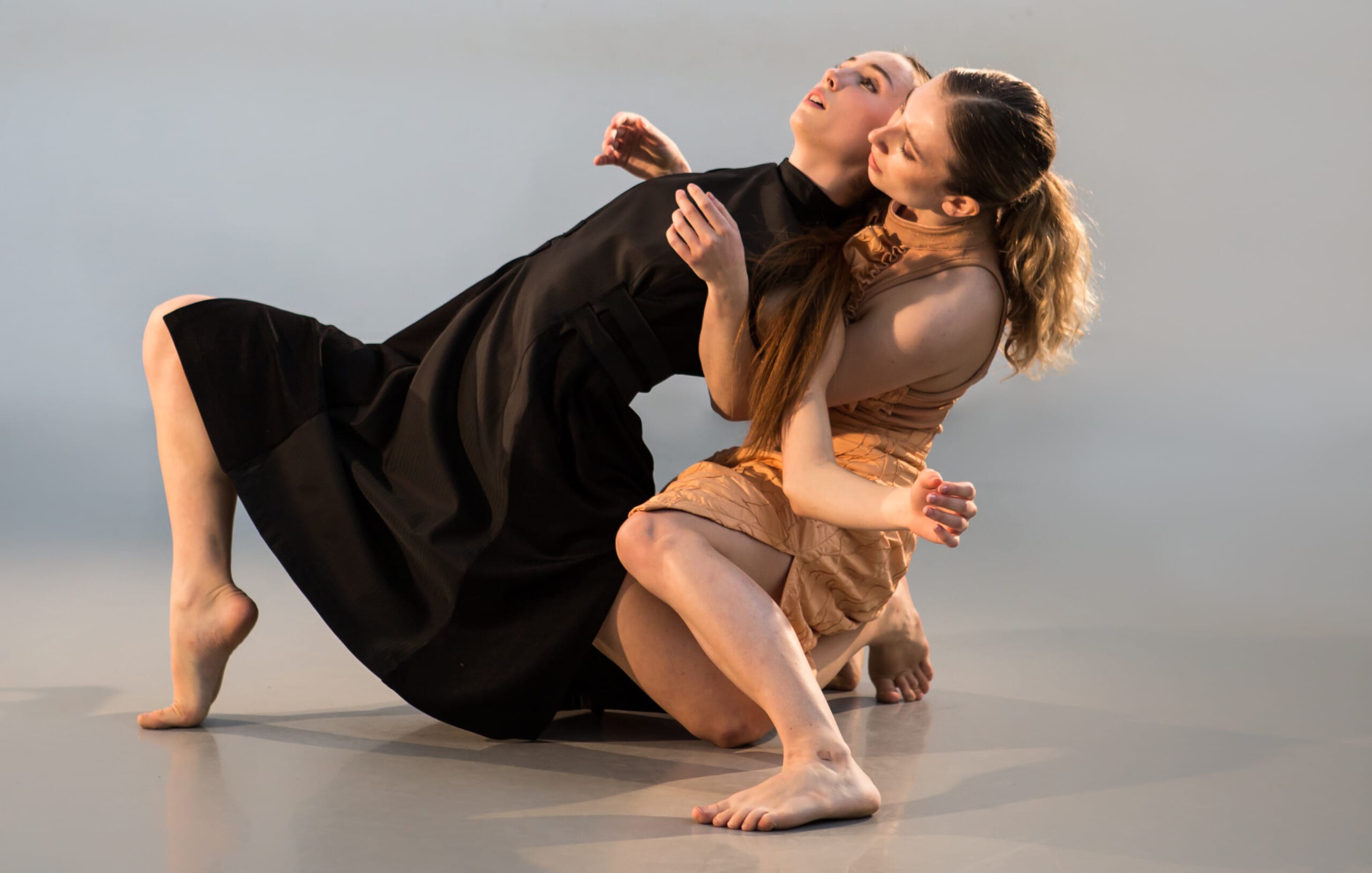 Two female dancers engage in floor work. The performer on the left wears a black dress and leans backwards. The performer on the right wears a nude dress, and supports her fellow performer in a duet pose.