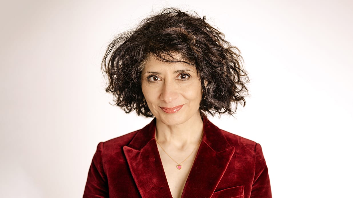 Comedian Shaparak Khorsandi pictured from the shoulders up. She is wearing a rust coloured velvet jacket and a necklace and has chin-length curly hair. She is slightly smiling at the camera.