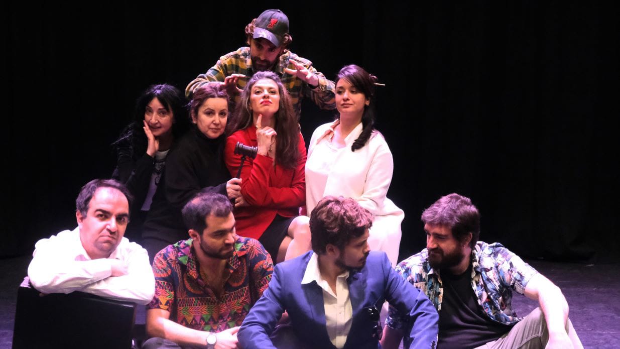 The cast of I Am A Lioness. Four cast members in the foreground are seated and looking at each other. On the second row, four women look at the viewer as unseen behind them, a man reaches forwards towards one of the characters.