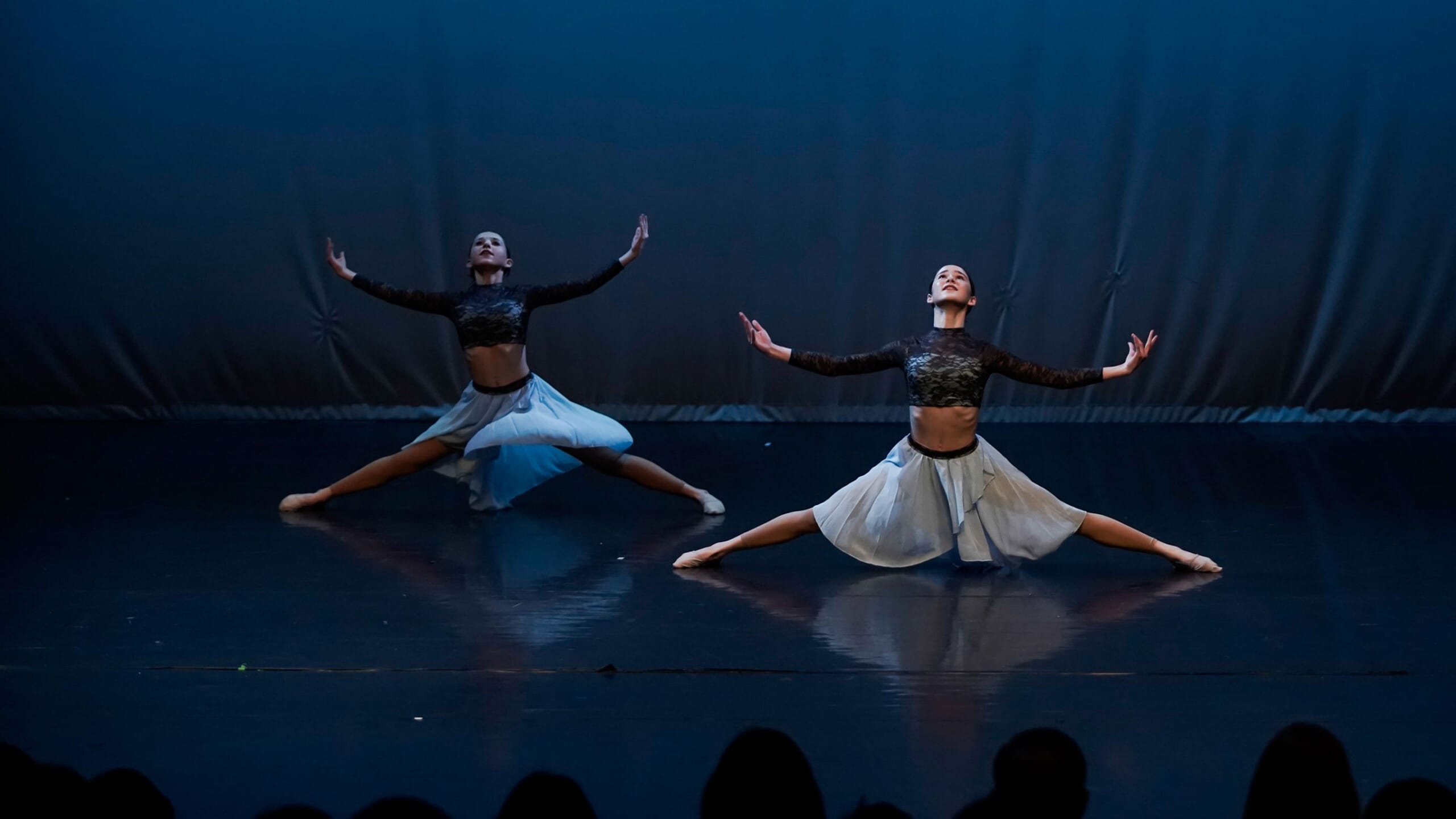 Two young dancers on a dark stage doing box splits with their arms stretched wide to their sides. In the foreground, the audience can be seen in silhouette.