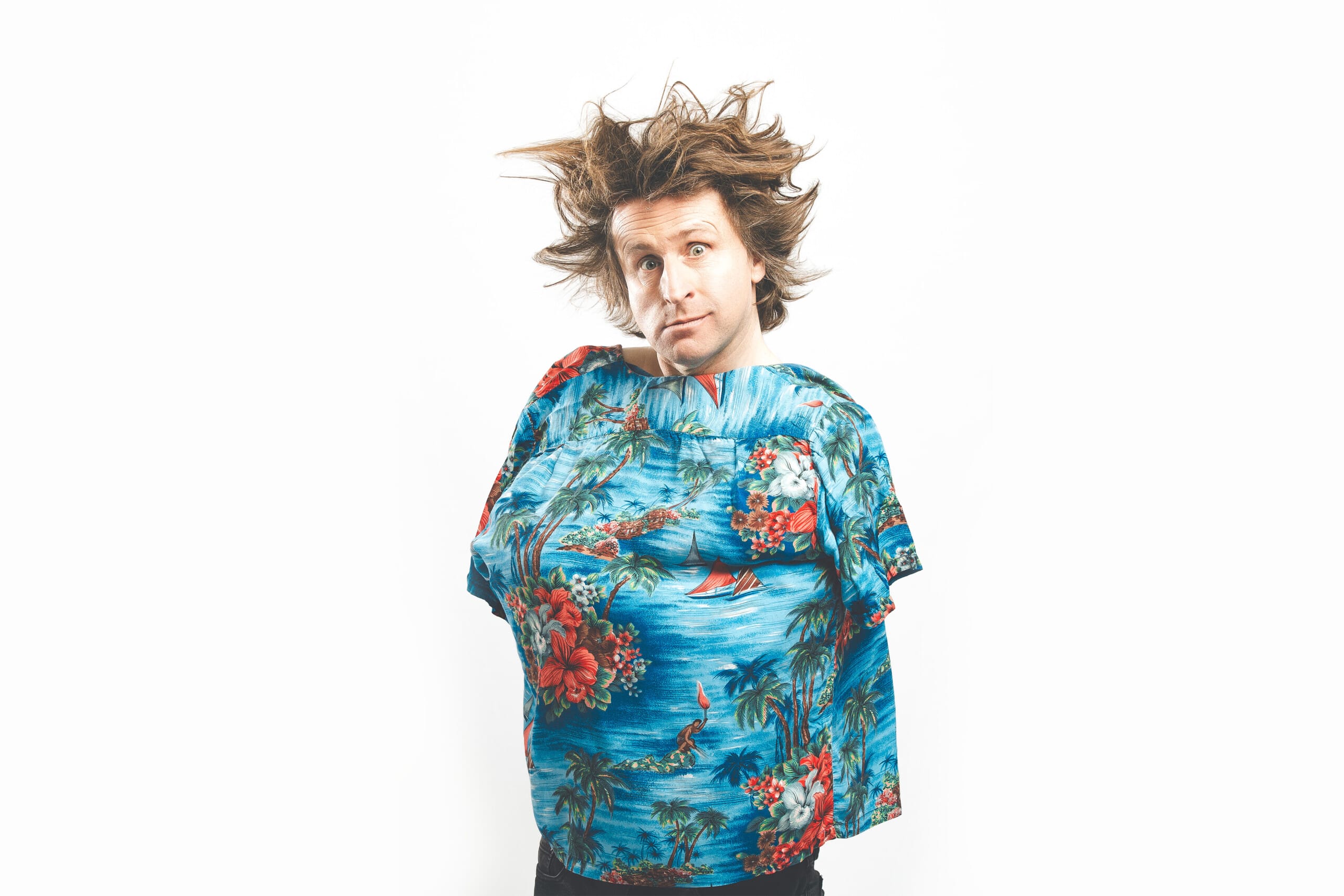 Comedian Milton Jones poses in a vibrant blue Hawaiian shirt, worn backwards. His hair is blown dramatically with a wind-machine, and he wears a bemused, comical expression.