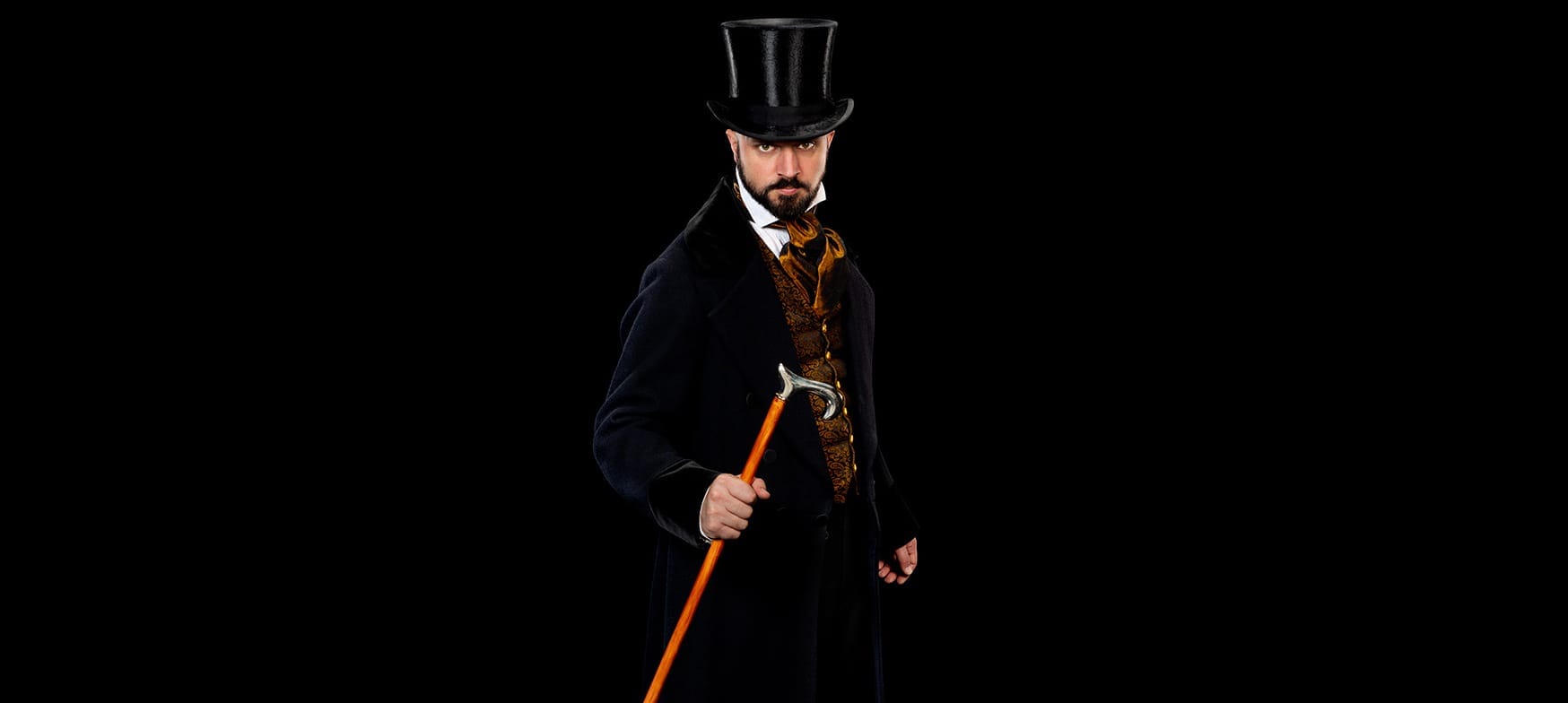 Andrew Margerison, the performer in A Christmas Carol, is wearing a Victorian outfit including a top hat, long dark jacket, and cane. He is staring at the viewer.