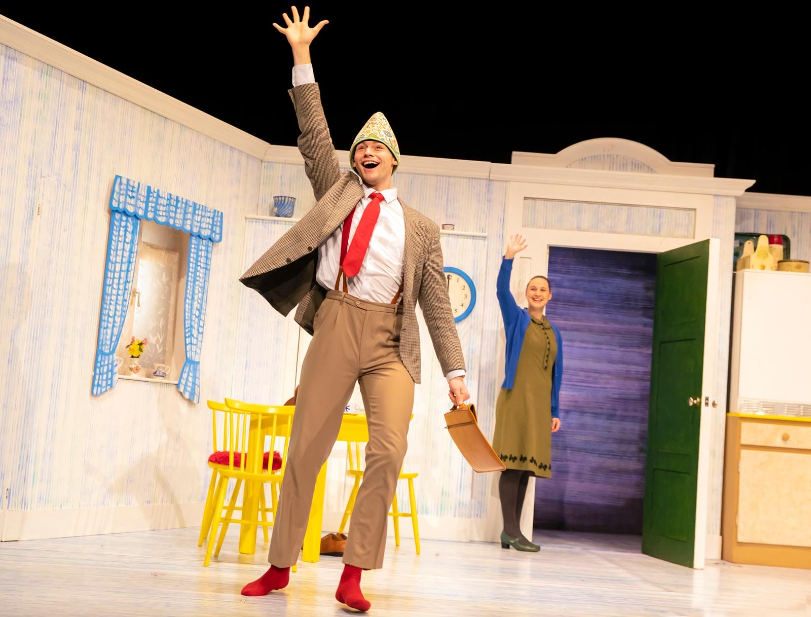 The character of Daddy on stage in The Tiger Who Came to Tea. He is in the foreground waving enthusiastically. The character of Mummy is in the background, waving by the open door.