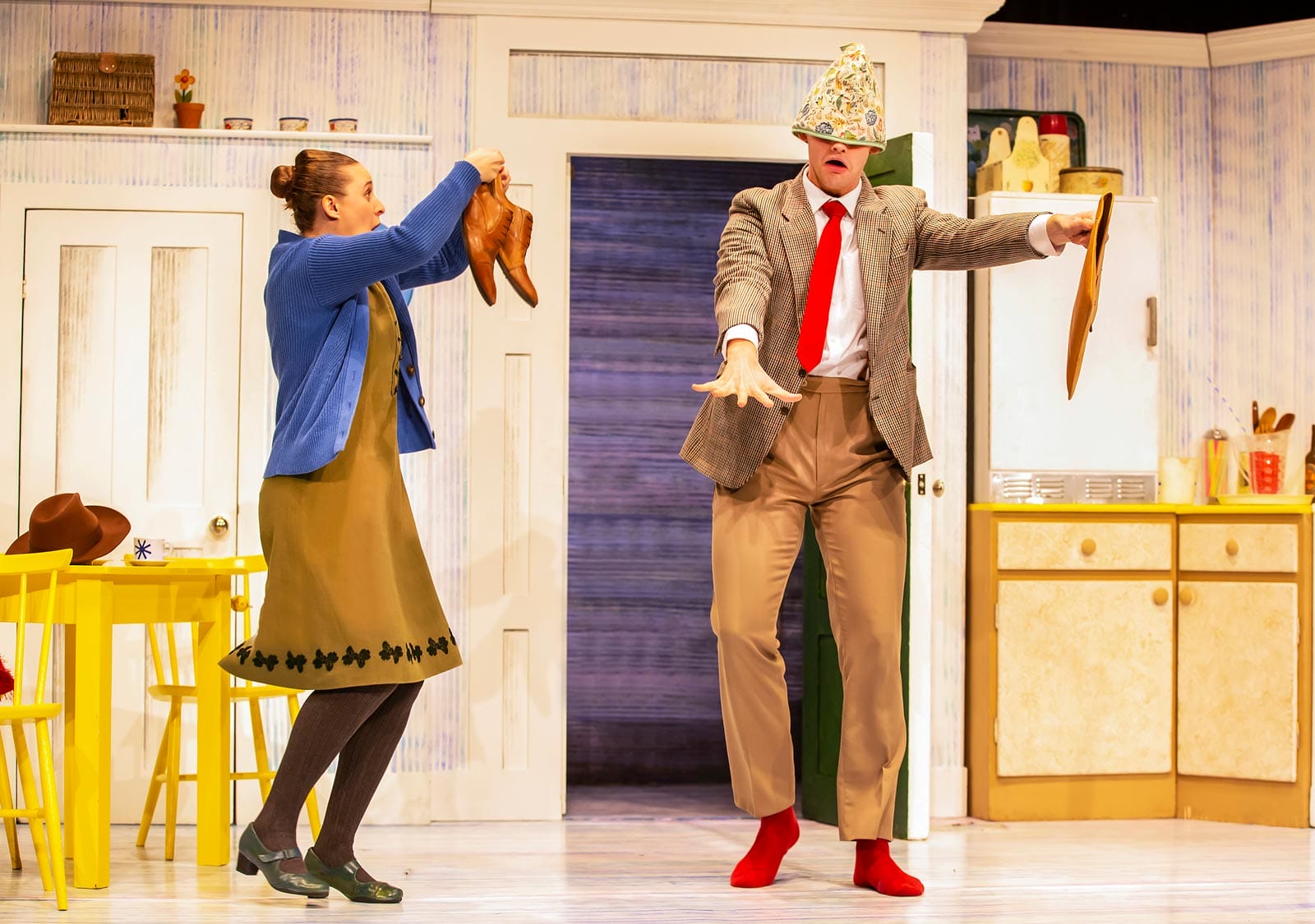 The characters of Mummy and Daddy on stage in The Tiger Who Came to Tea. Daddy has a tea cosy on his head covering his eyes and his arms stretched out in front of him. Mummy holds up his shoes.