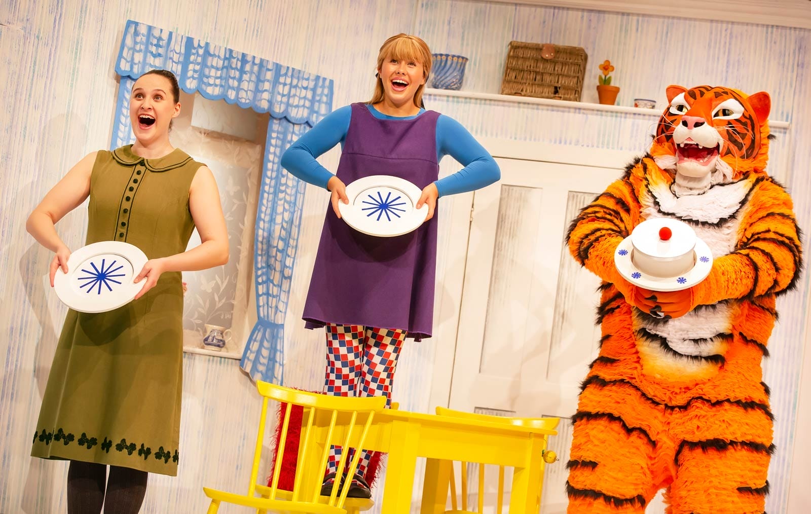Performers playing Mummy and Sophie in The Tiger Who Came to Tea are holding empty plates, The Tiger holds a plate with a large white cake on it.