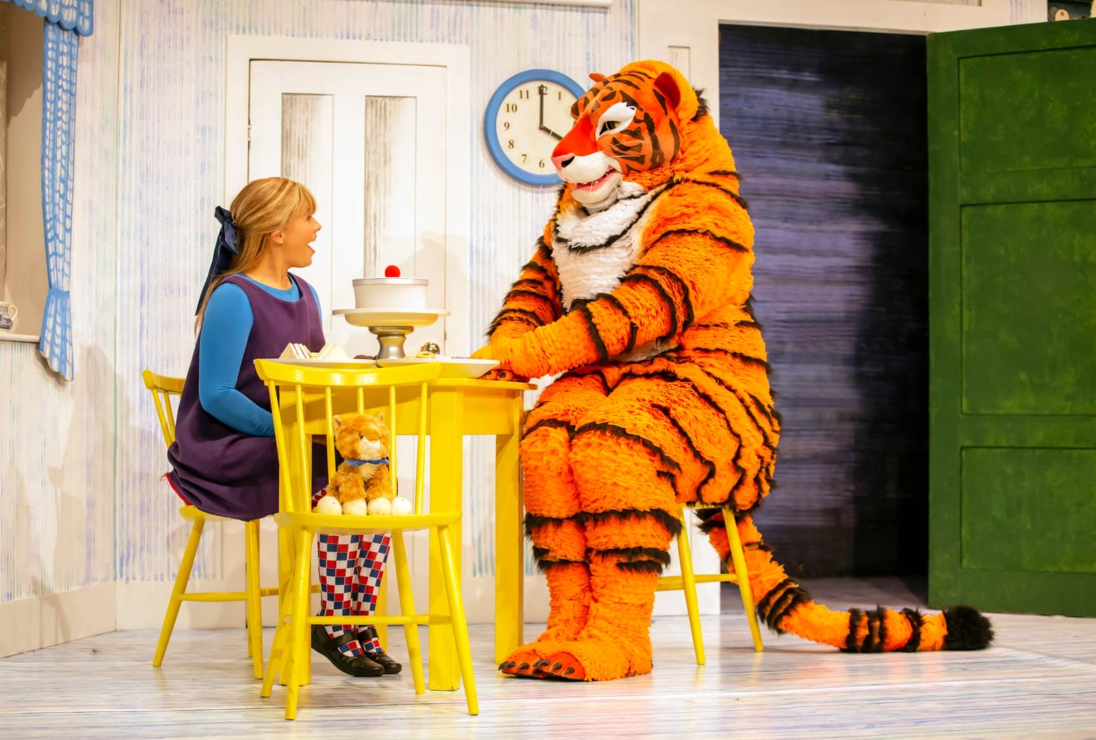 Performers playing Sophie and the Tiger in The Tiger Who Came to Tea are sat at a yellow painted table. A large cake with a cherry on top is between them.
