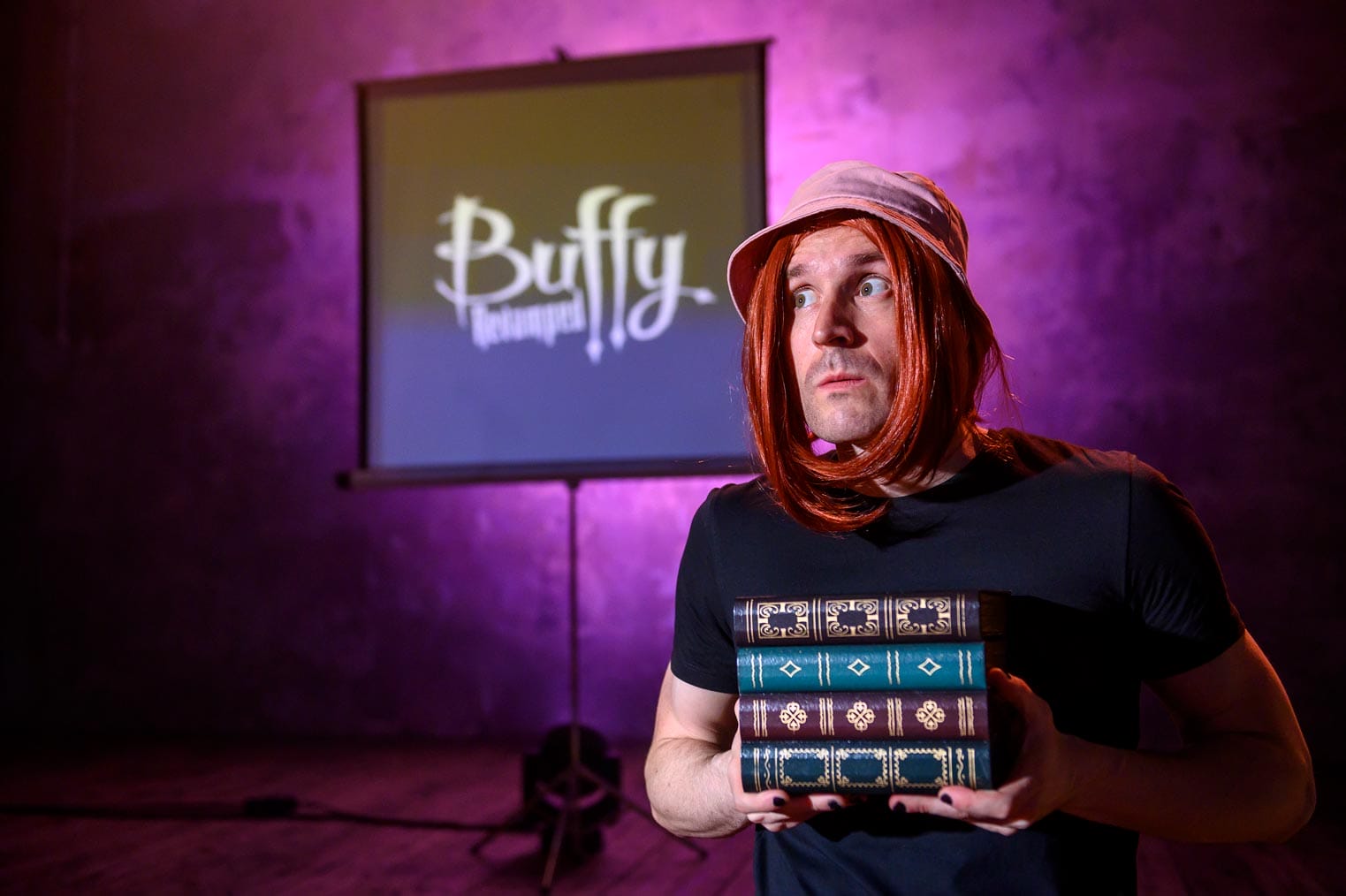 The Spike character holding holy books wearing an orange wig and a hat with the text Buffy in the backdrop projected on a screen.