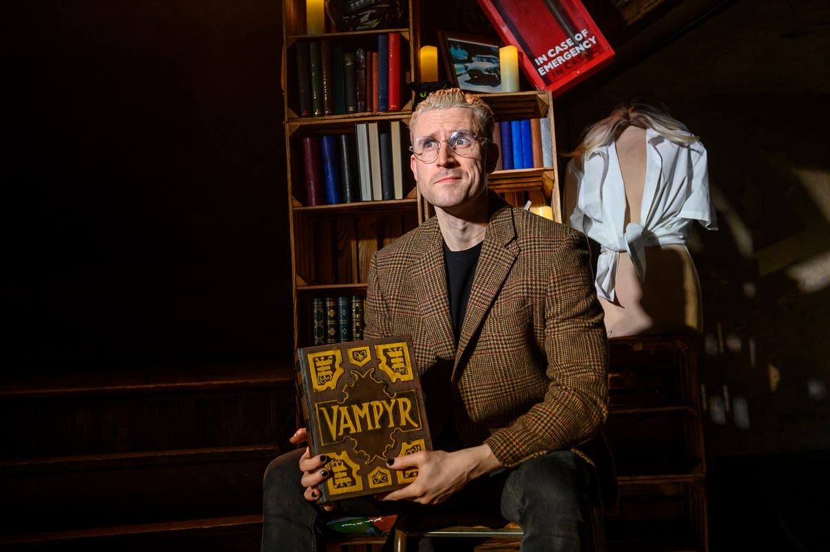 The character Spike sitting on a chair holding a Vampire book in a suit, with a disorganised bookshelf in the background.