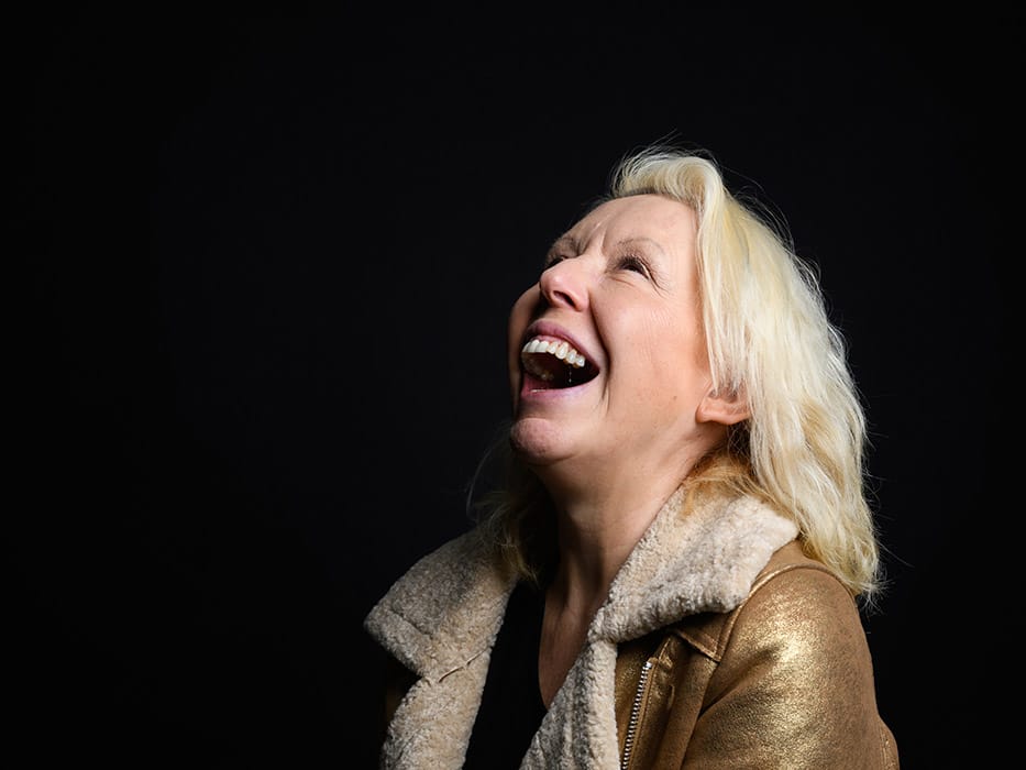 An image of Barb Jungr laughing out loud in a beig jacket in front of a black back drop.