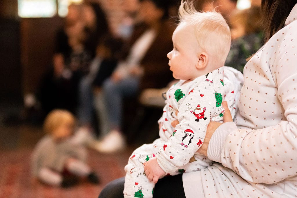 An image of a baby in a Christmas onesie sitting on the knees of an adult watching the show in awe.
