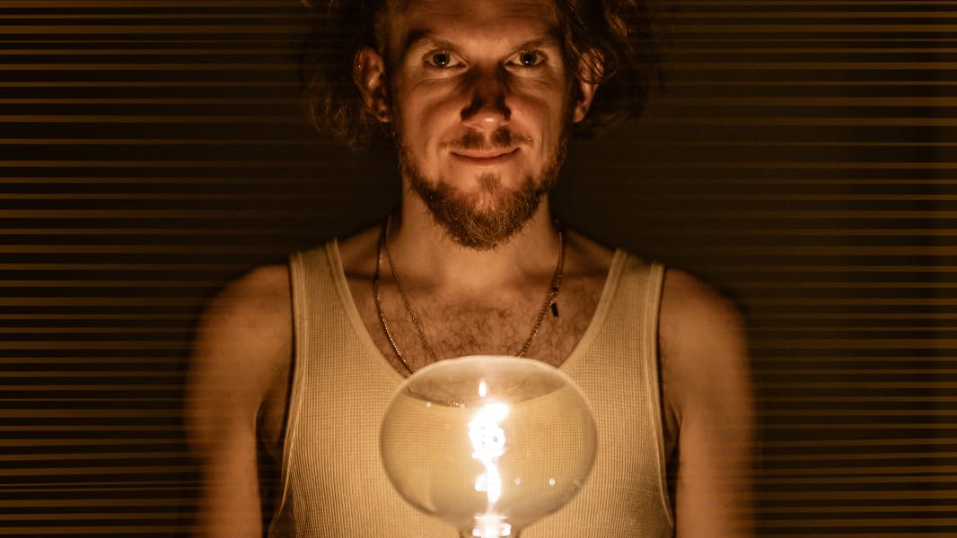 Picture of a man wearing a white vest and necklaces, looking at the camera. He is holding a large lightbulb which illuminates his face. He is surrounded by darkness.