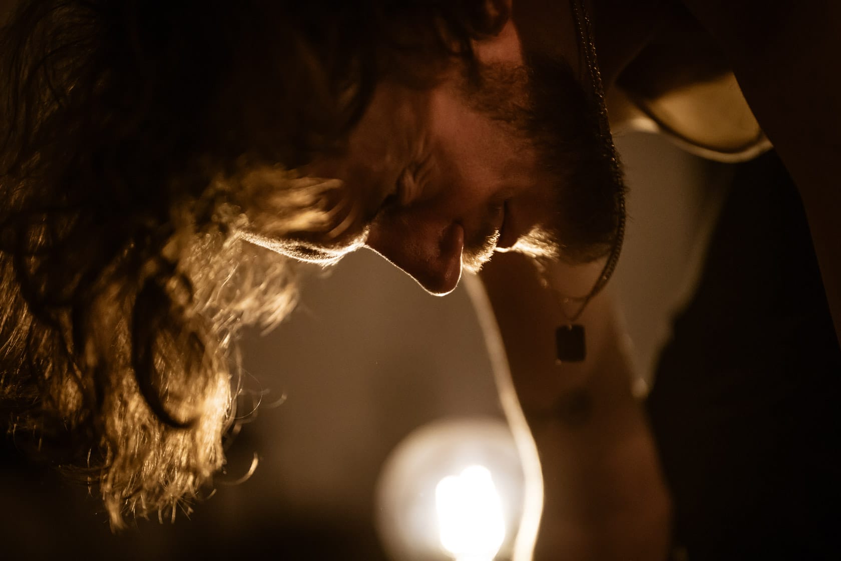 Image is a close up of a performer leaning down. His long hair is lit by a large lightbulb which can be seen in the background.