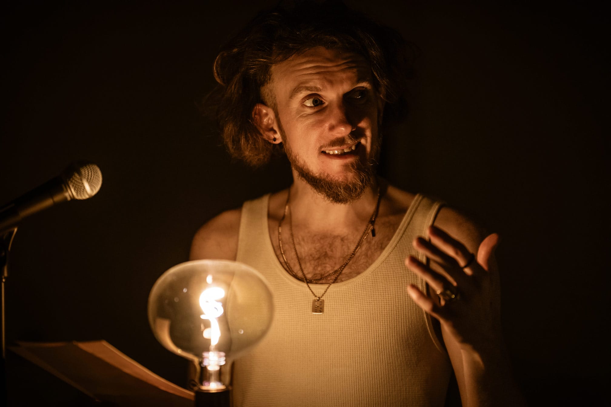 Image of a performer on stage, he is a young man wearing a white vest and necklaces. He is surrounded by darkness but illuminated by a large lightbulb in front of him. He is holding a piece of paper in one hand and gesturing with the other. A microphone can be seen next to him.