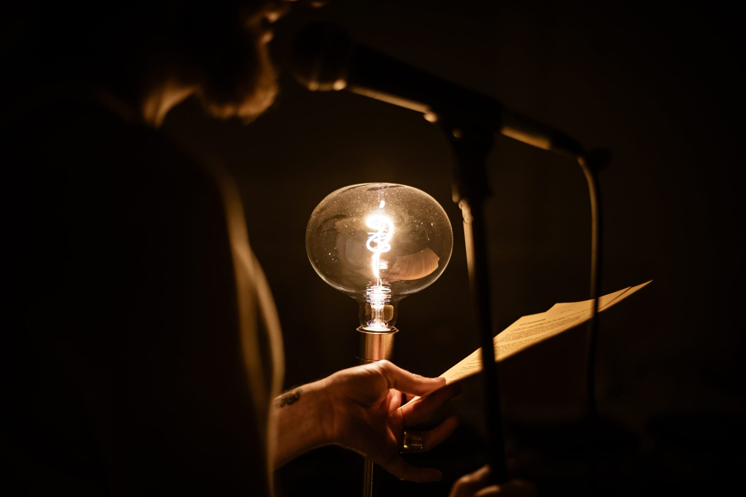 Close up of the shoulders and lower face of a performer on stage. They are standing in front of a microphone and surrounded by darkness and lit by a large lightbulb.