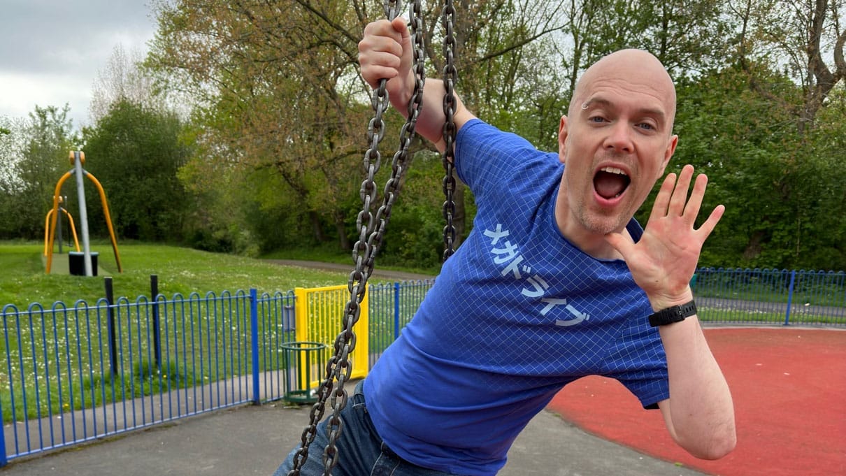 Poet Dommy B is pictured outside in a children's playground. He is holding onto the chains of a swing with one hand and the other is next to his mouth as he shouts.