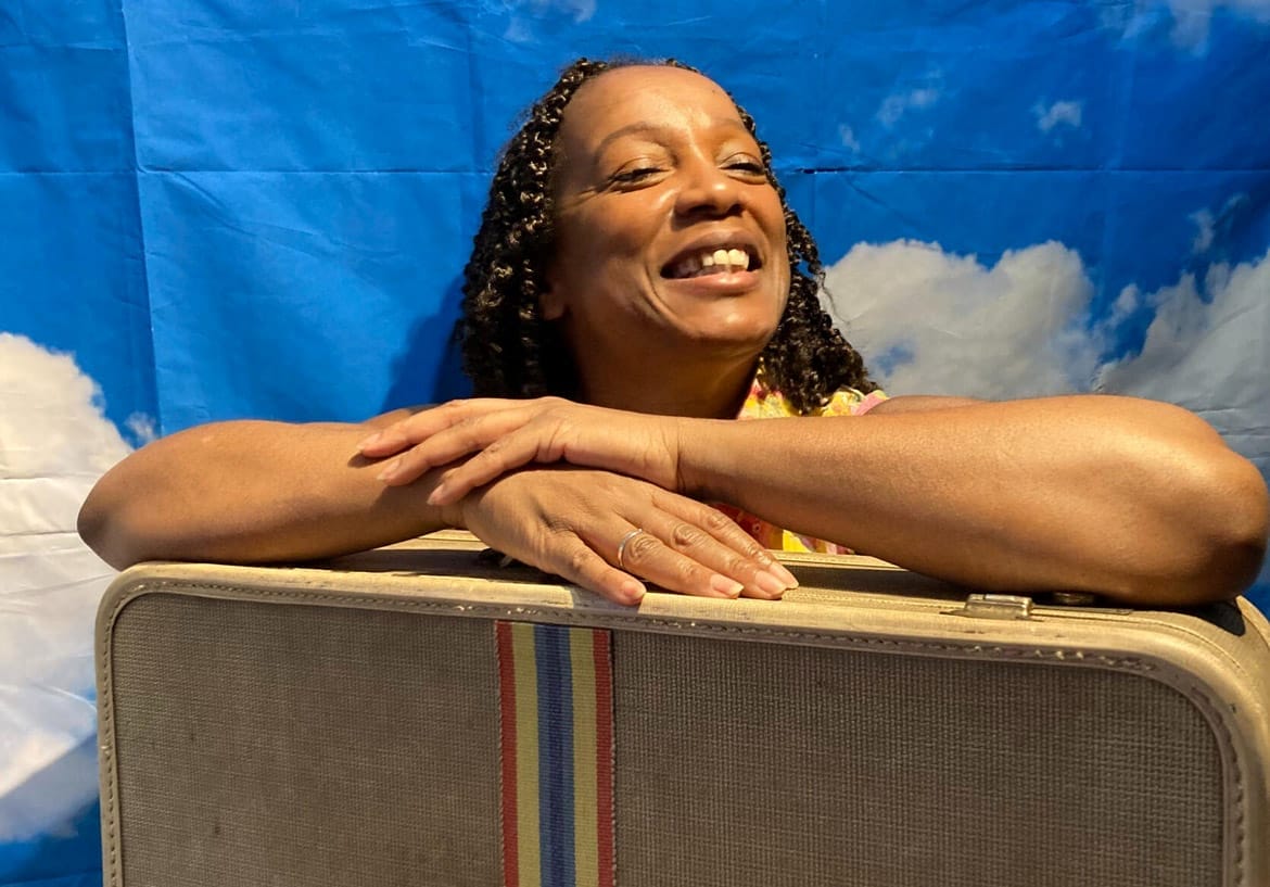 A woman smiles with her eyes closed and her hands crossed over a suitcase in front of her. Behind her is a background of a bright blue sky with fluffy white clouds.