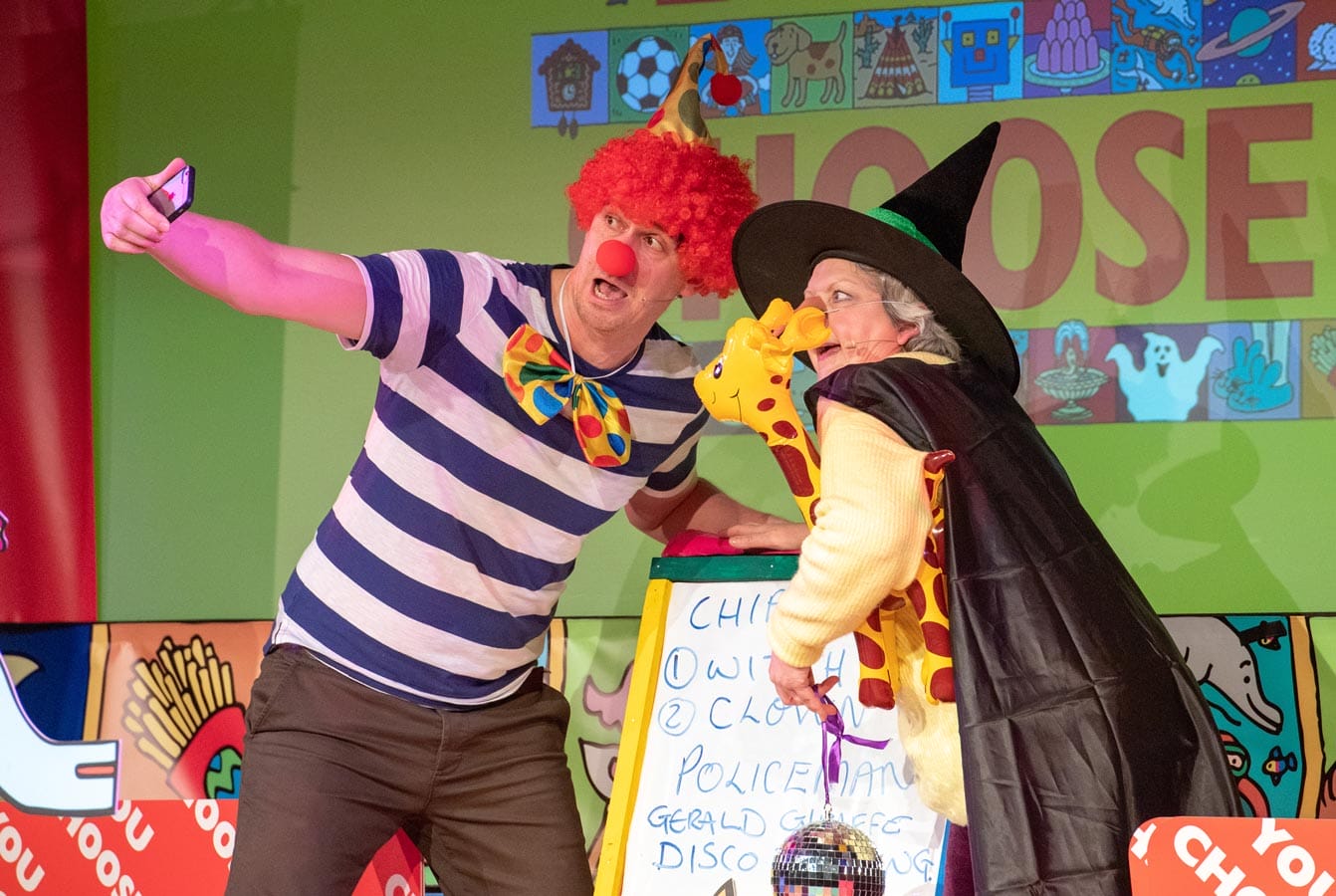 A person dressed as a clown with a red wig, clown nose, and colorful bow tie takes a selfie with another person dressed as a witch, holding a giraffe toy. They are on stage in front of a green backdrop with colorful illustrations.