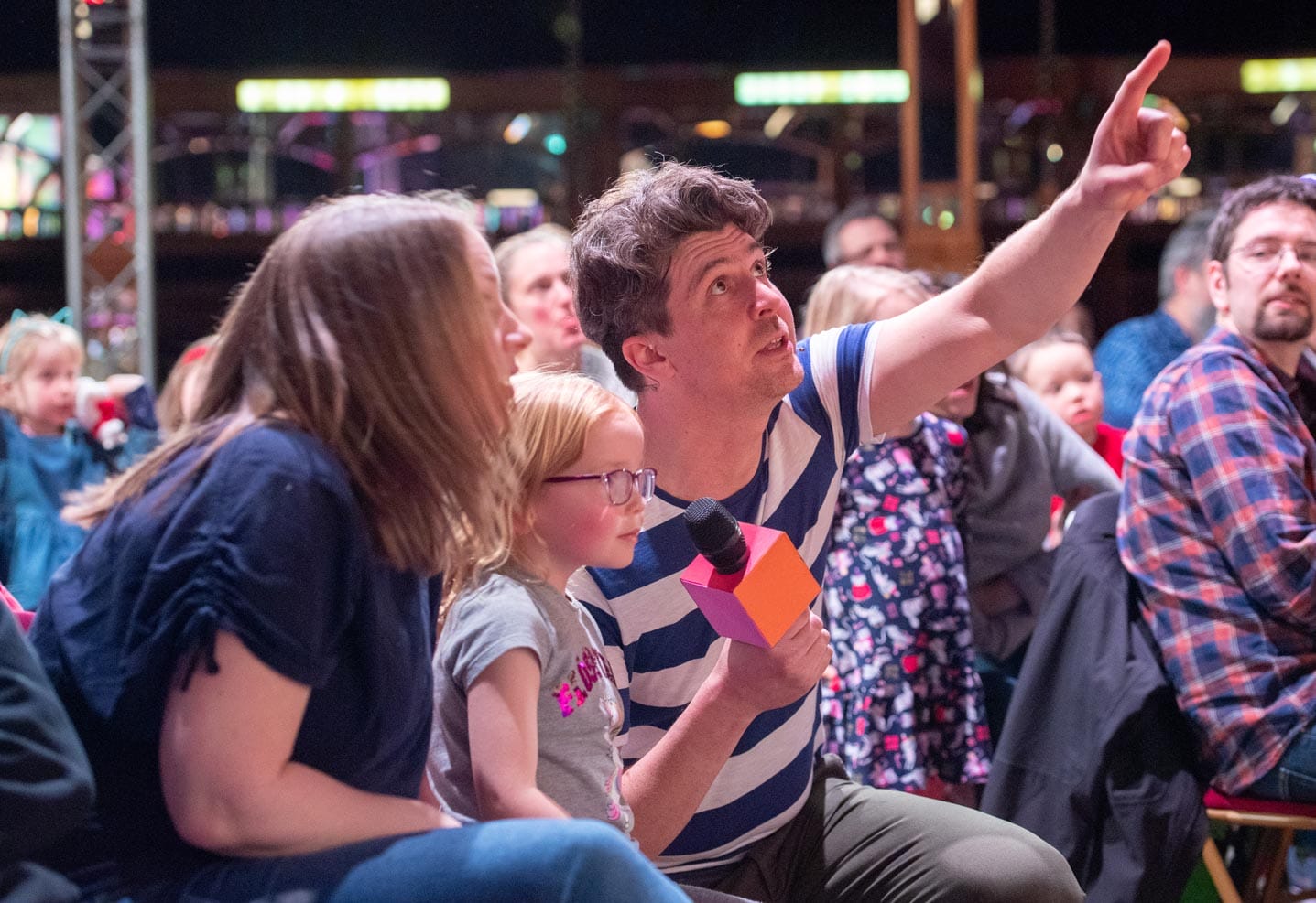 A man in a striped shirt holds a microphone and points upwards while sitting among a group of children and adults in an audience. A woman and a young girl wearing glasses are seated next to him, looking in the direction he's pointing.