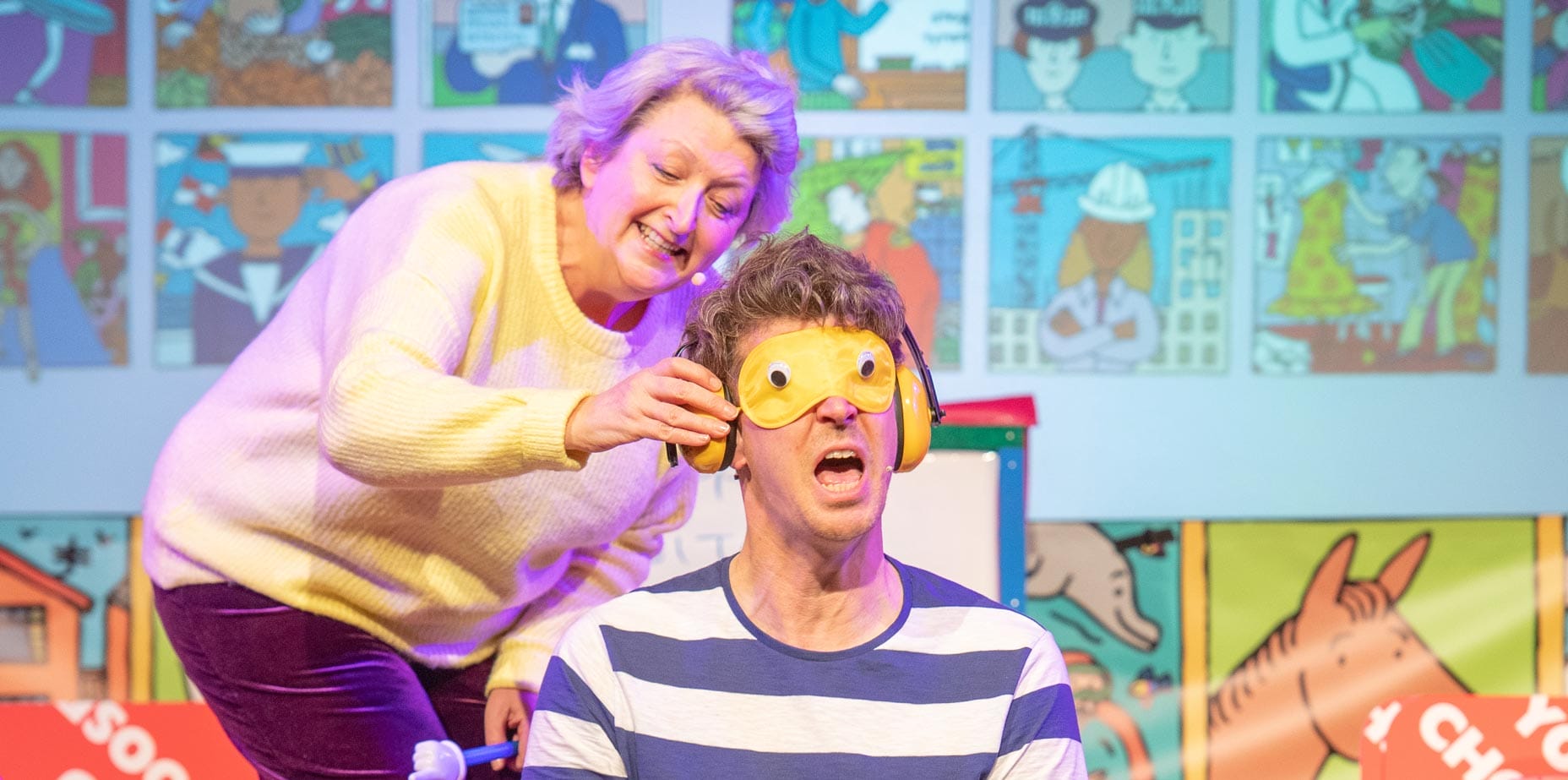 The female performer in yellow is placing an ear defender on the male performer's ears and a yellow sleeping eye mask on his eyes.