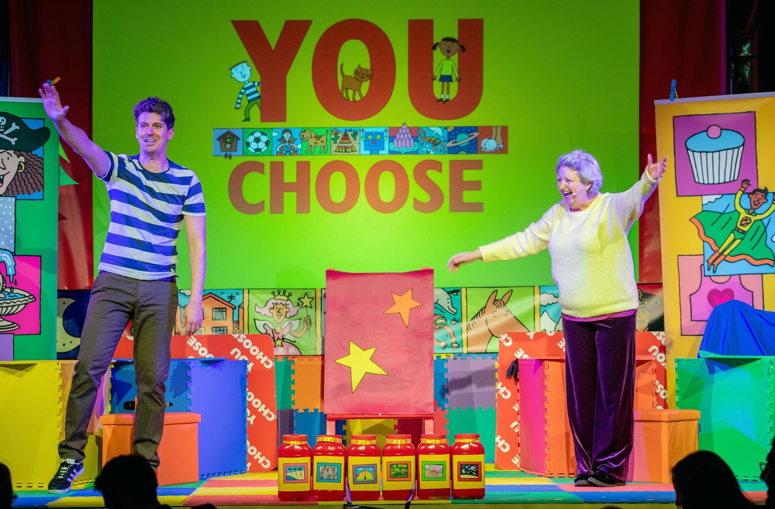Two performers, a man and a woman, are on stage waving to the audience. The backdrop reads