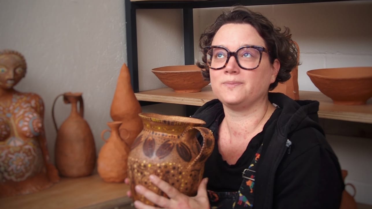 Potter Amy Wilson is pictured in her studio. She is holding a terracotta pot with a handle, behind her are other pots, bowls and vessels on shelves and a table.