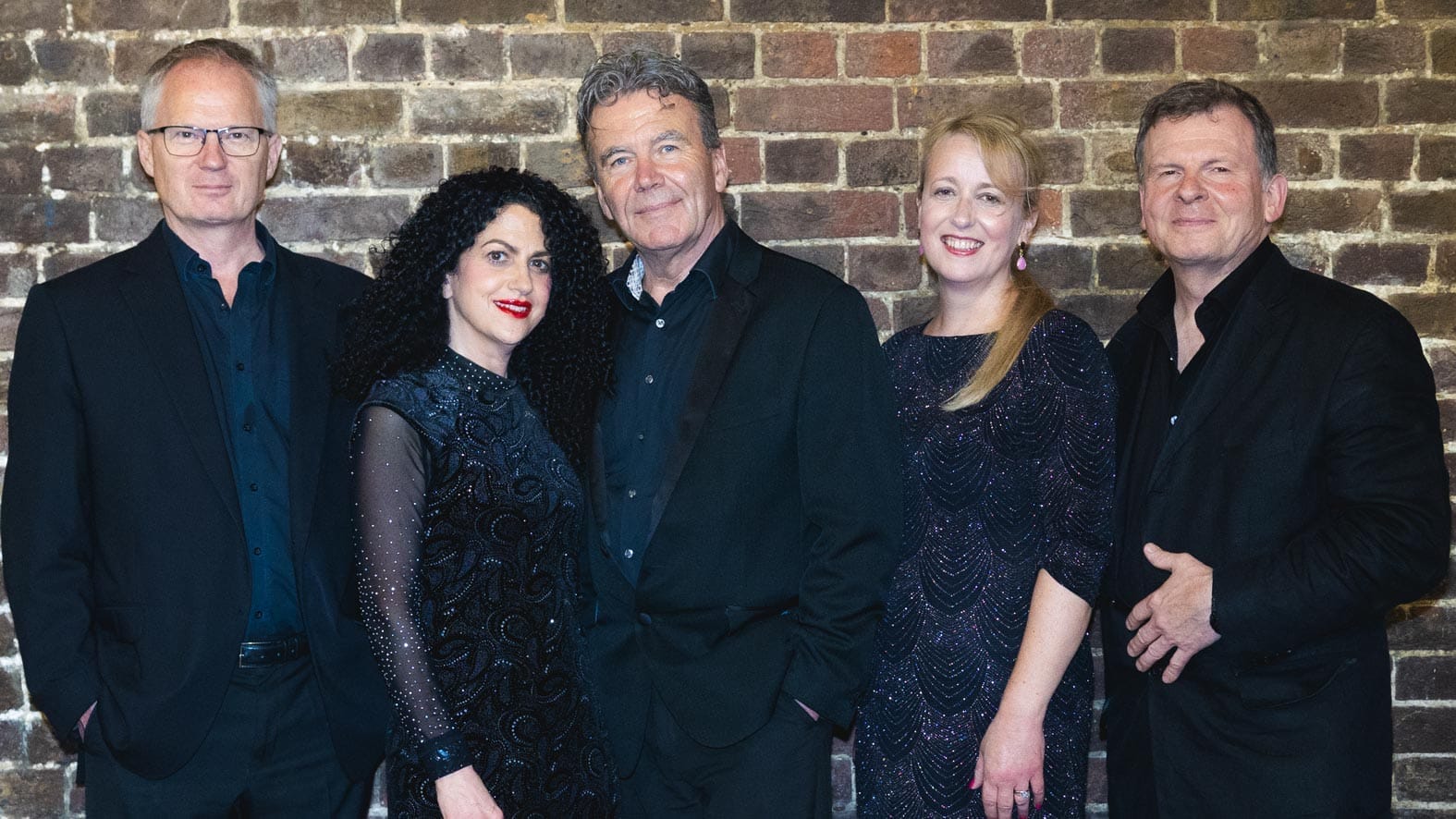 The five members of Sabra Swing wearing smart black clothes are pictured in front of a brick wall.