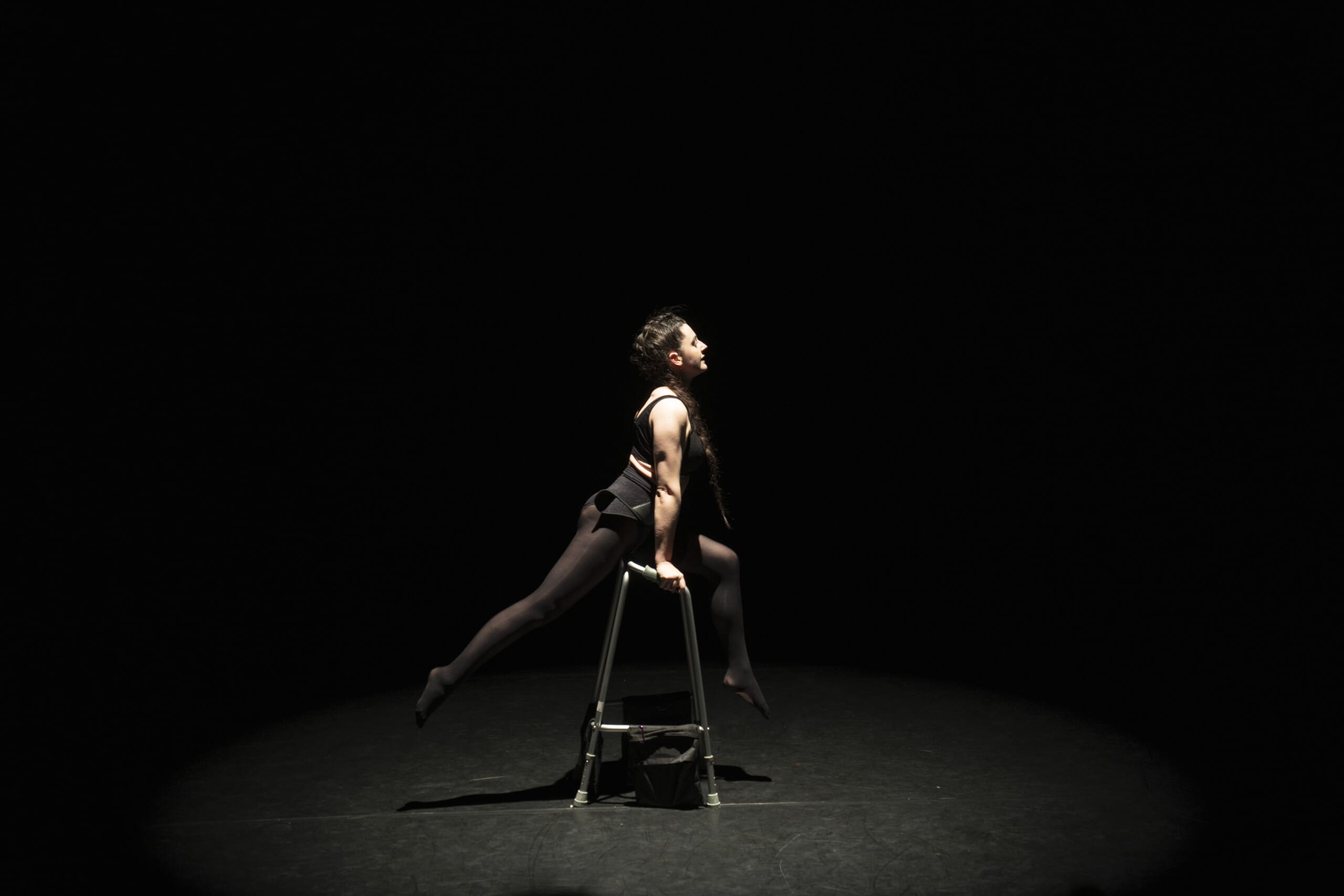 Porcelain a white woman with dark hair tied in a braid is wearing black tights, shorts, a hip brace and crop top. She is performing on stage and is balancing on a zimmer frame with one bent leg resting over the top bar, her hands holding on at each side to provide extra support and her other leg extended behind her pointing away from her frame.