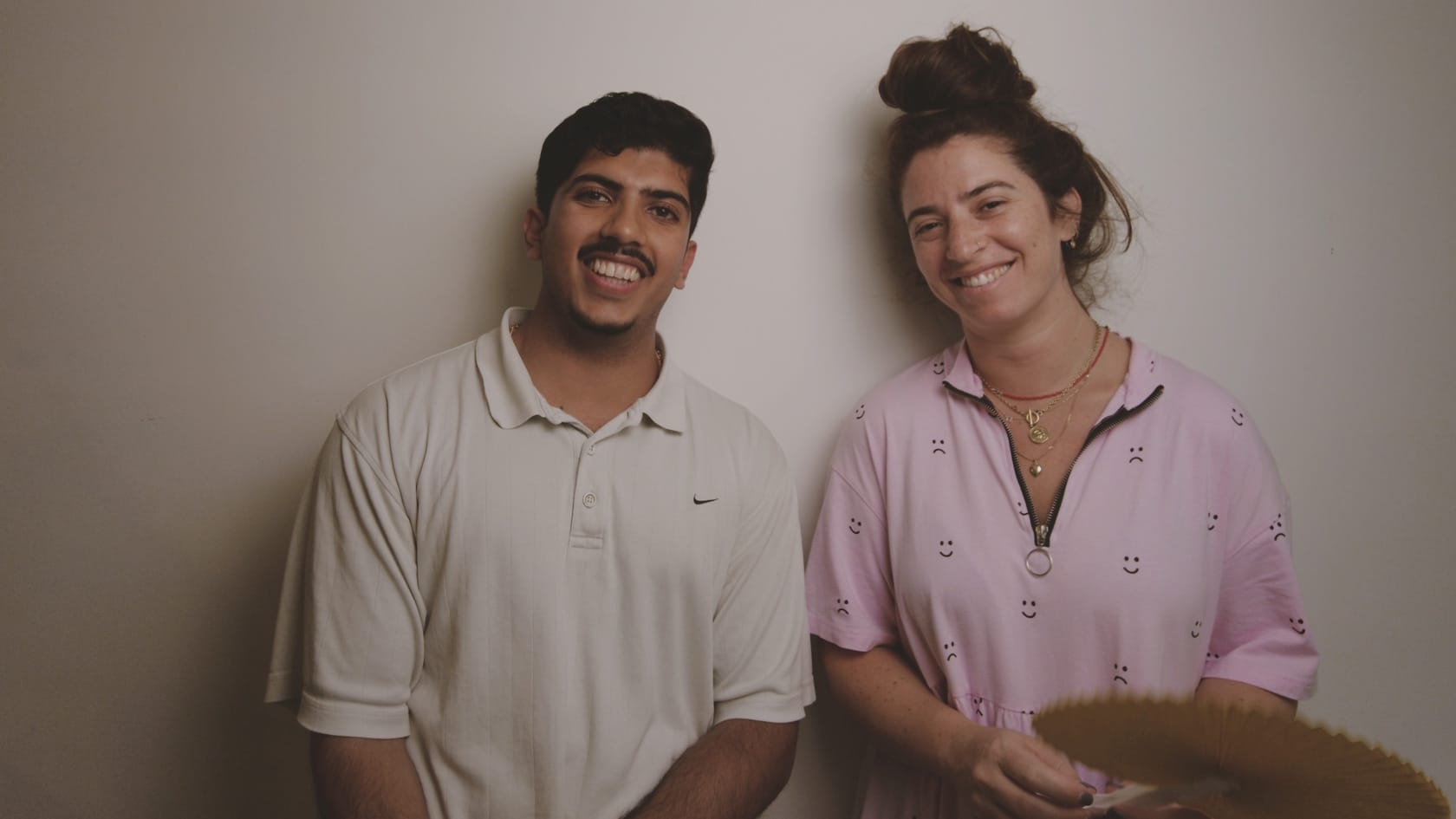 Adi Gortler and Hamza Ali sit with their backs against a white wall, smiling at the viewer.