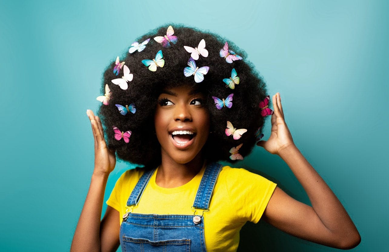 An image of Playworker Amber wearing a bright yellow t-shirt and denim dungarees, her afro hair is covered in colourful butterflies