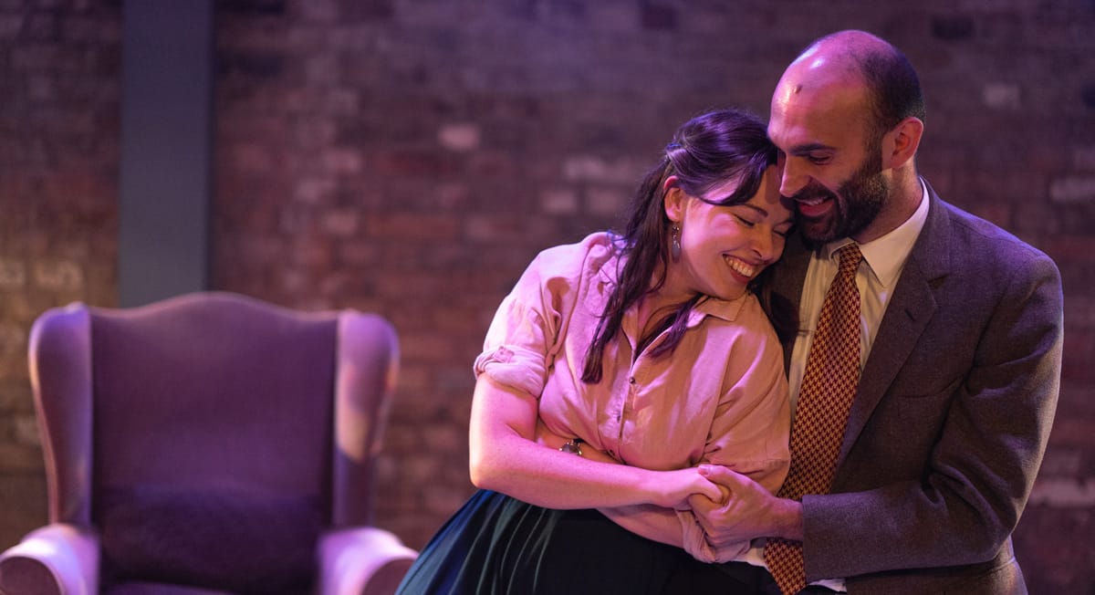 A brunette woman in white shirt leans into the arms of a man in suit.