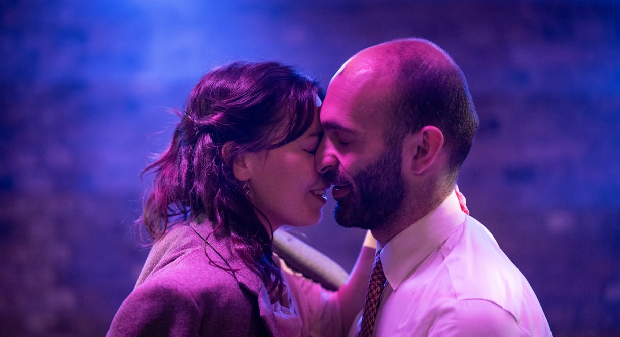 A bearded man and a brunette woman share an intimate moment embracing one another.