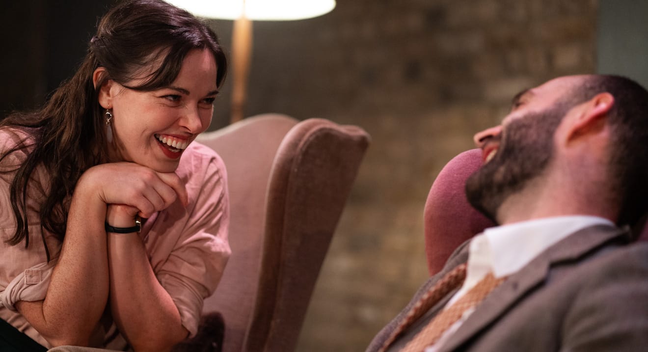 A brunette woman giggling whilst looking at a bearded man talking to her.