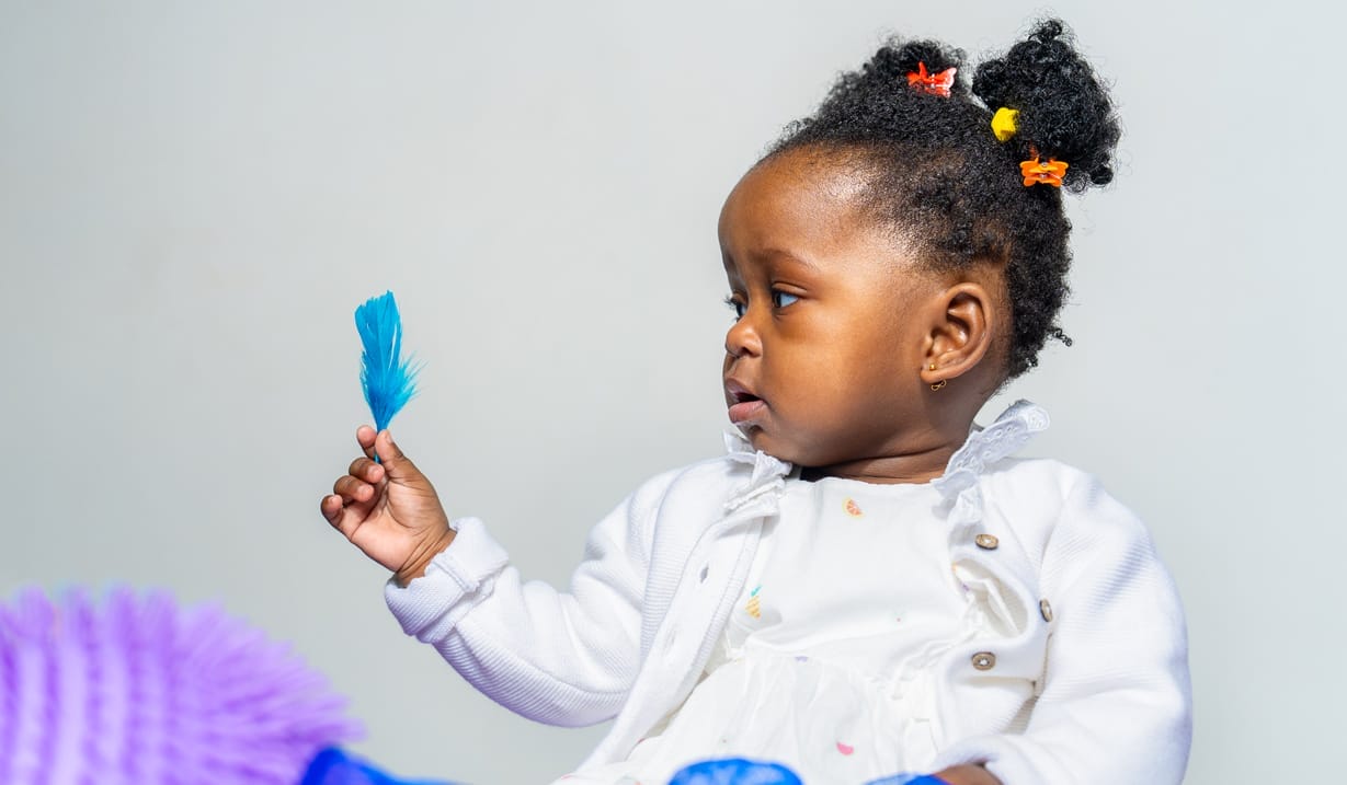 A baby holds a blue feather and is surrounded by textured fabrics.
