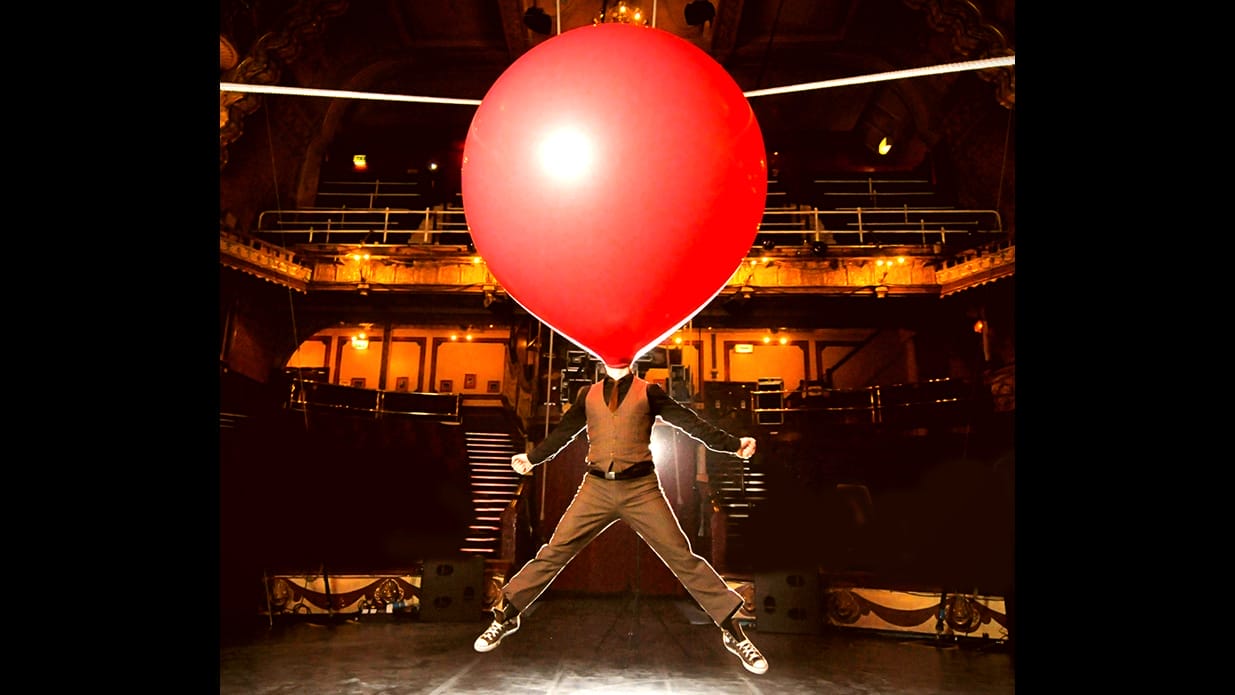 A performer on stage is doing a star jump with an enormous red balloon on their head.