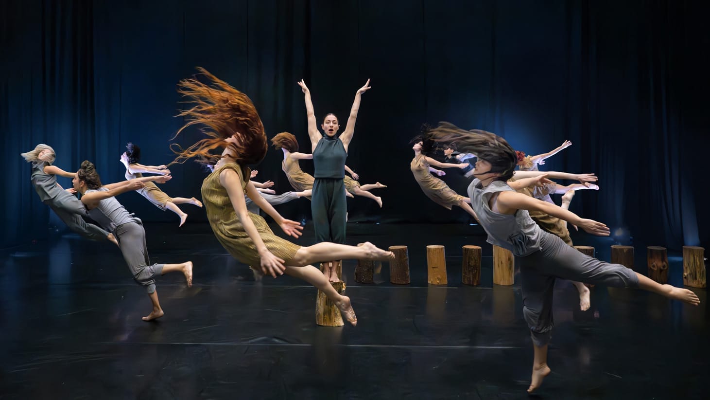 A group of dancers on stage, one stands in the middle on a block of wood with her arms raised, 10 others leap across the stage with their hair flying.
