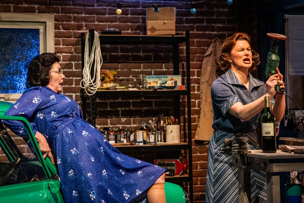 Production photo, one woman lying on a green car, the other holding a drill