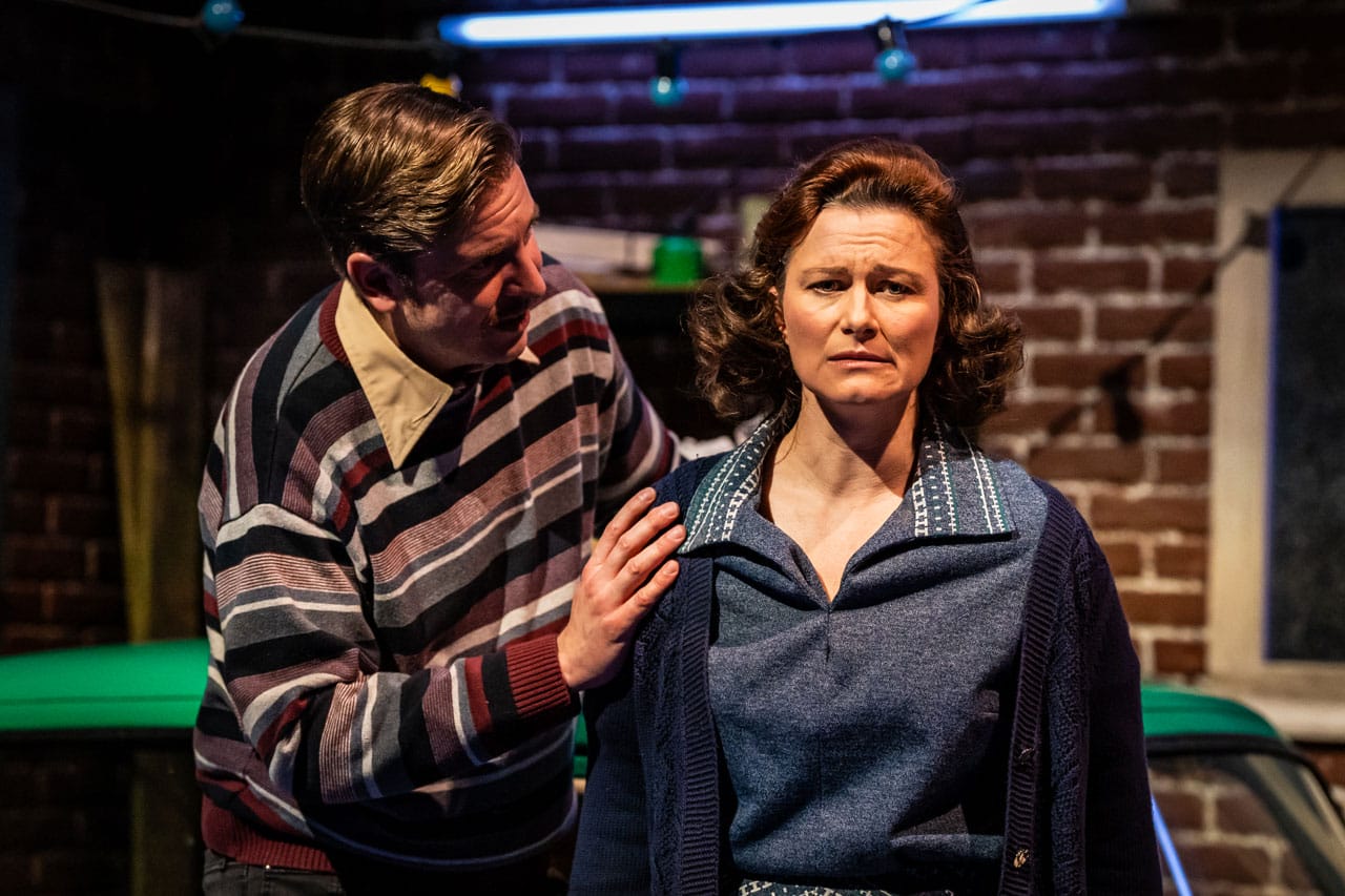 Production Photo. A woman looking at the audience and a man looking at her.