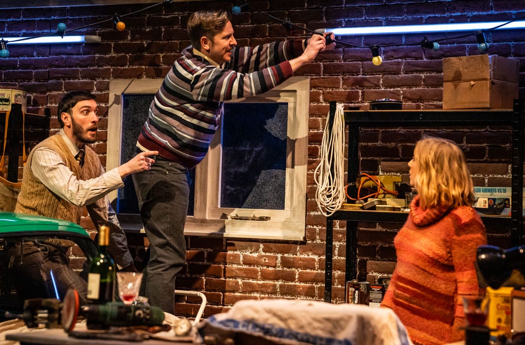 Production photo where one actor is putting up festoon lighting and two other actors stand around him.