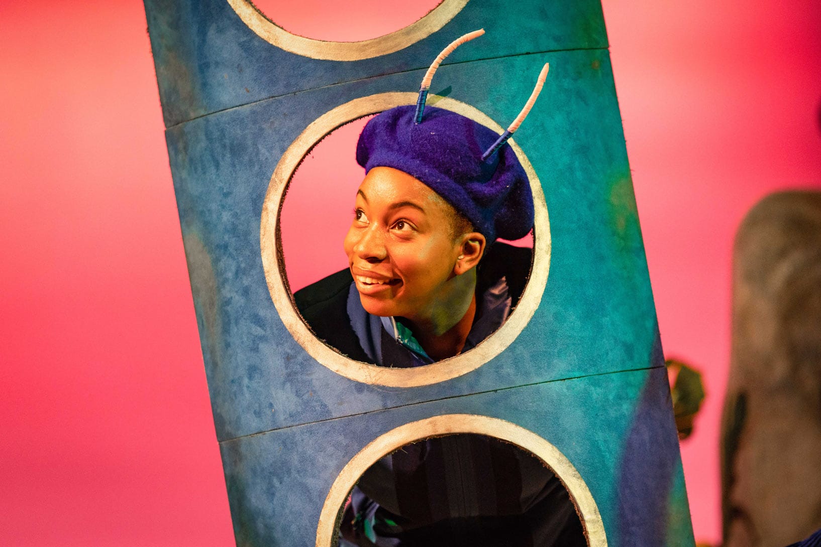 Felicia Akin-Tayo as Janet from The Smeds and Smoos smiles as she peeks through a circular prop against a pink background.