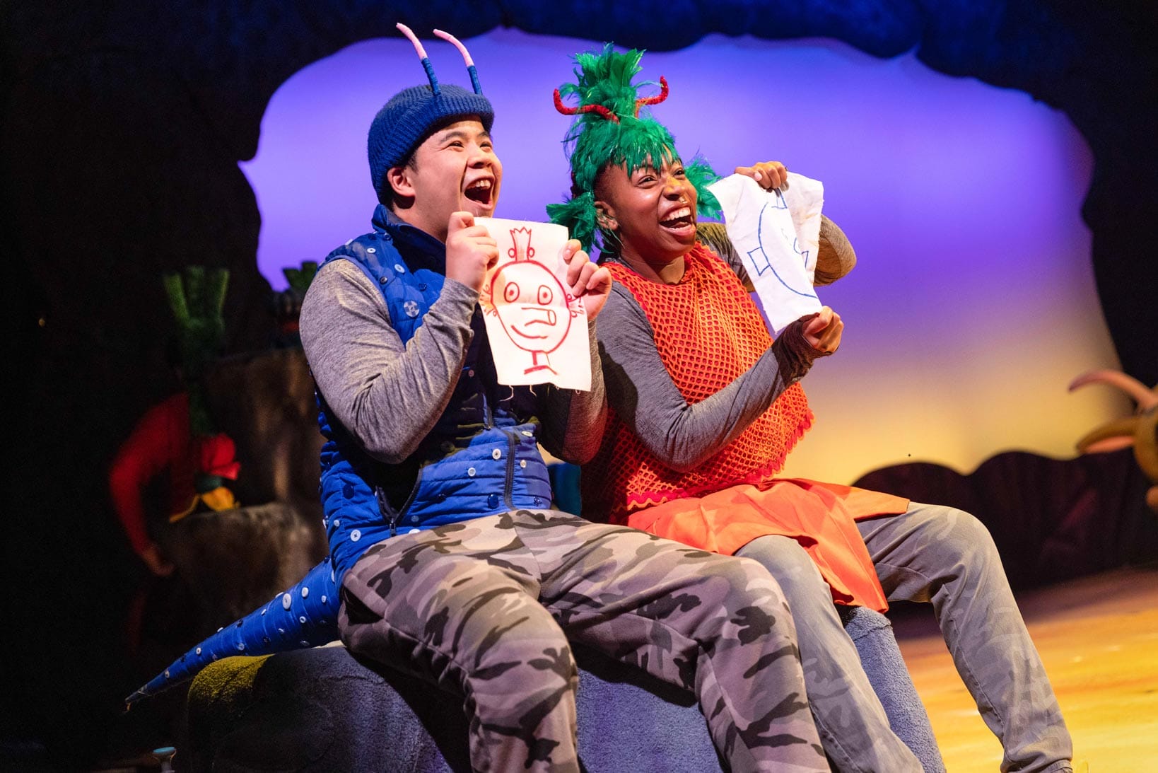 Robert Penny as Bill and Felicia Akin-Tayo as Janet from The Smeds and Smoos laugh while holding hand-drawn pictures on stage.